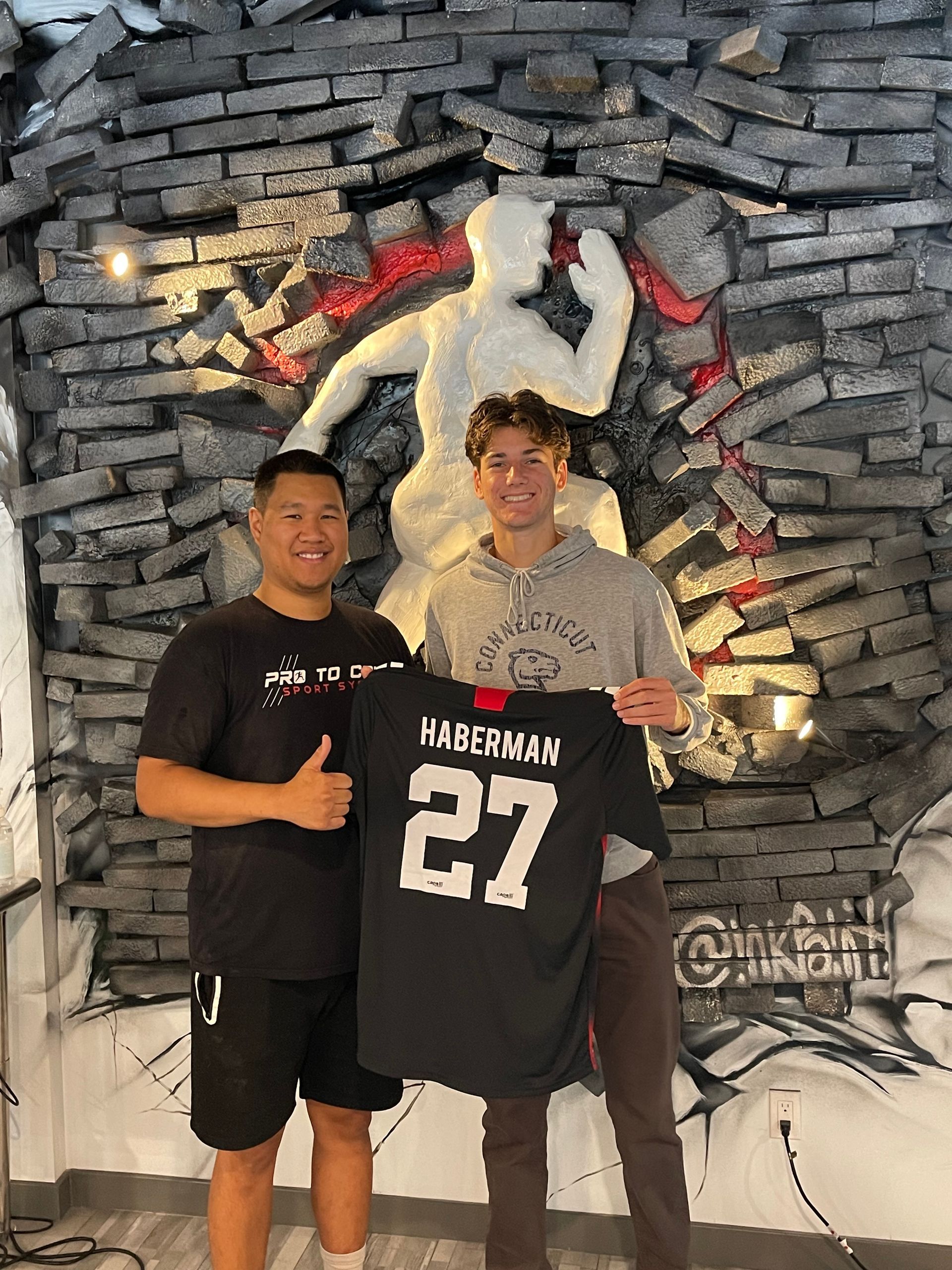 Two people posing with a black Haliburton #27 jersey in front of a basketball mural