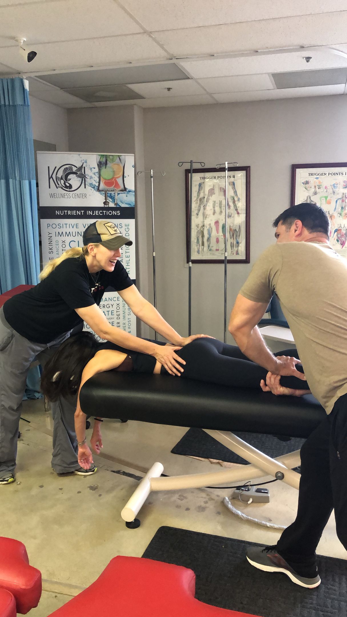 Two people moving a black padded table in a clinic room with posters on the walls.