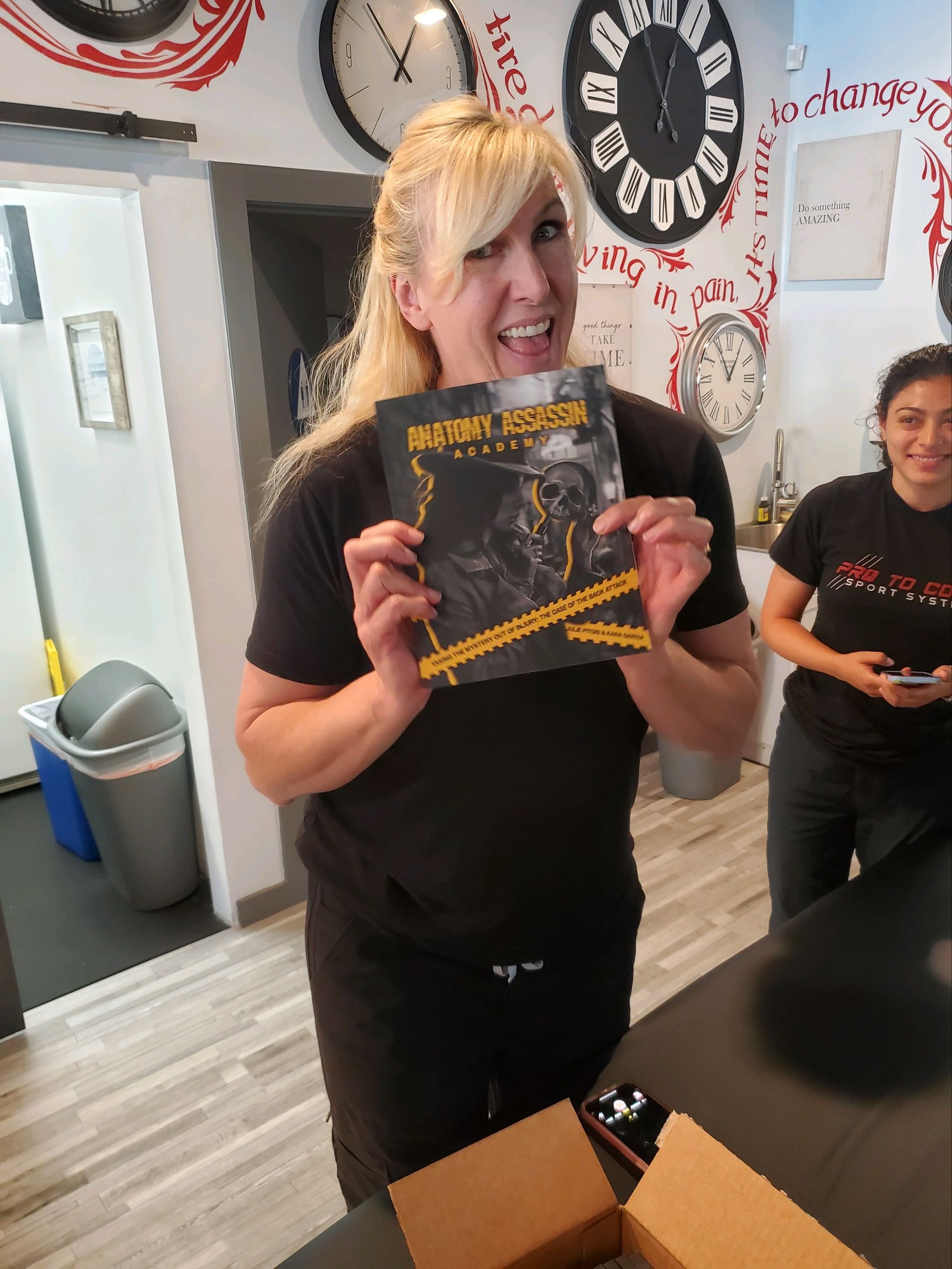 Woman smiling and holding a black-and-gold book in a bright room with wall clocks and another person nearby