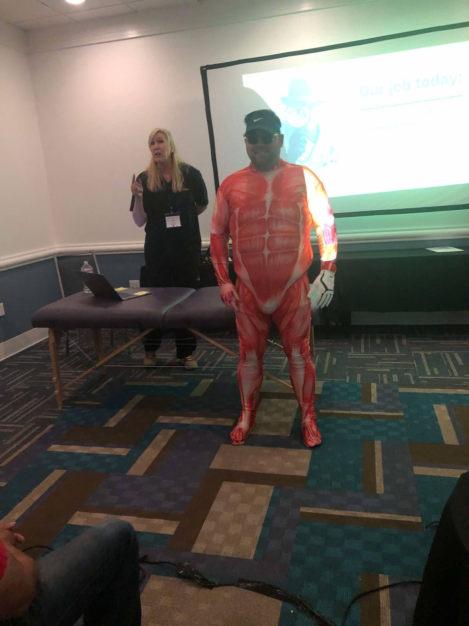 Person in a red bodysuit and cap stands beside a presenter in a conference room.