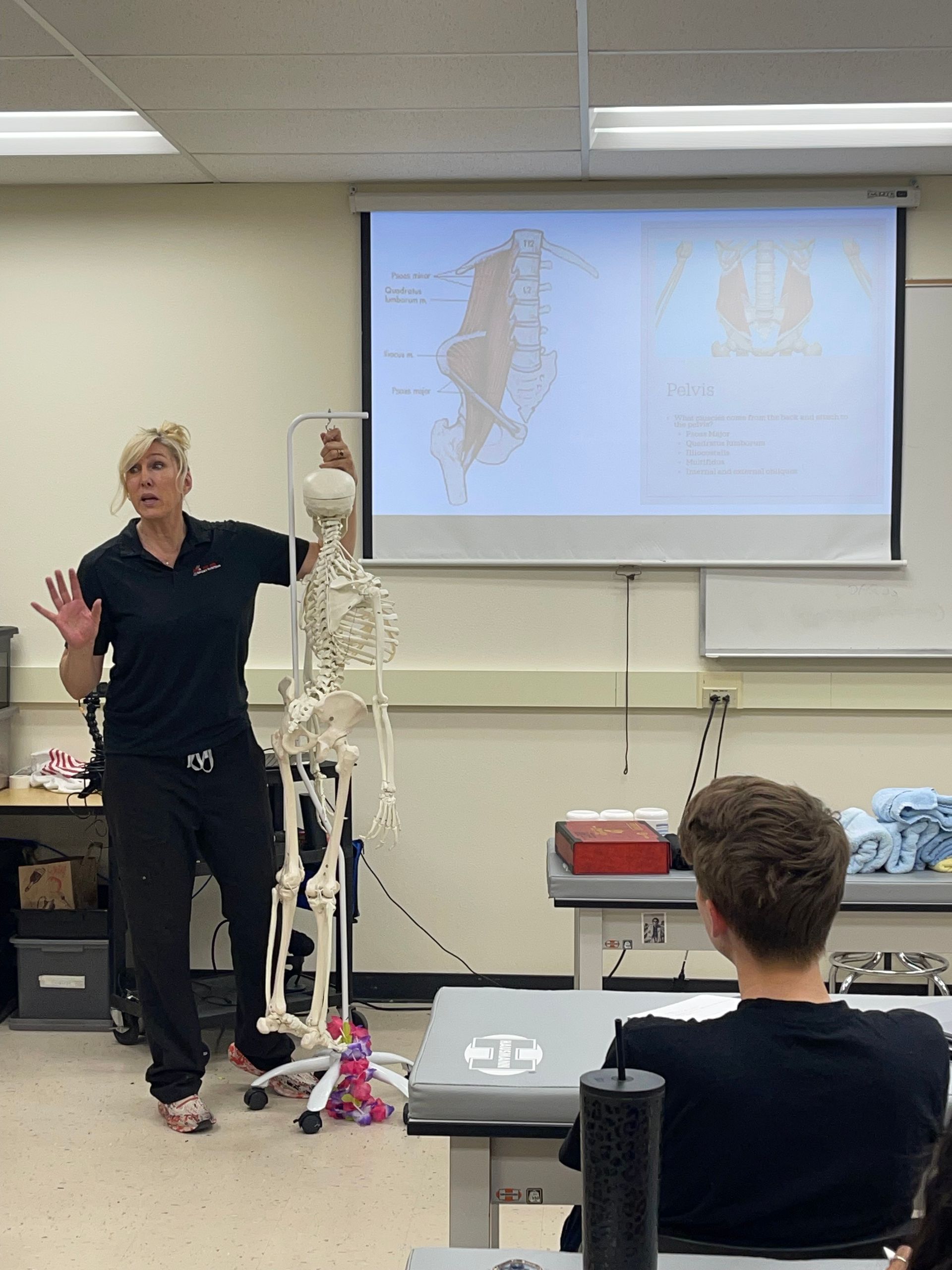 Instructor beside anatomical skeleton model, gesturing in a classroom with a projected spine diagram.