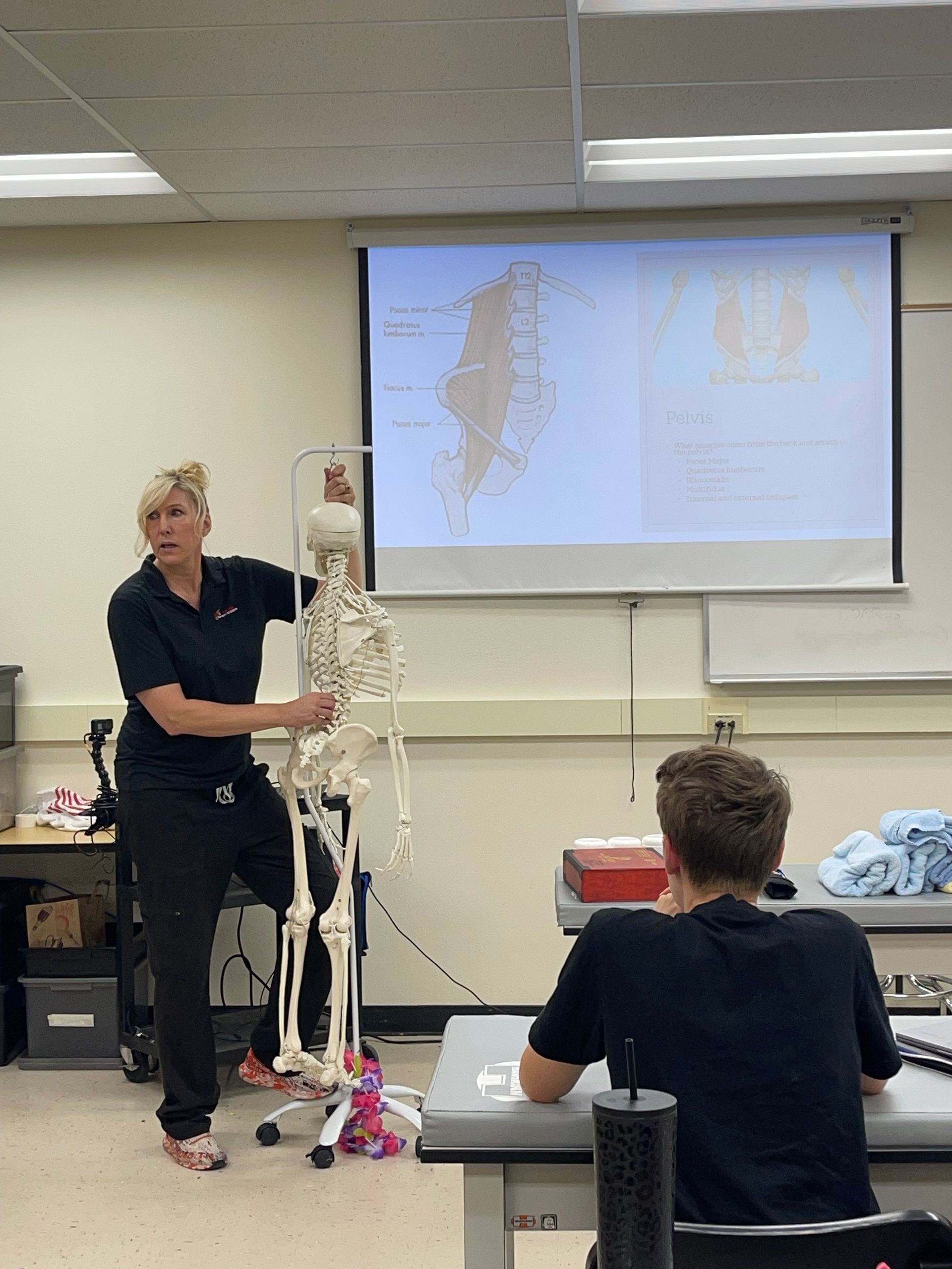 Instructor beside skeleton model teaching in classroom, with students seated and a projection screen behind her