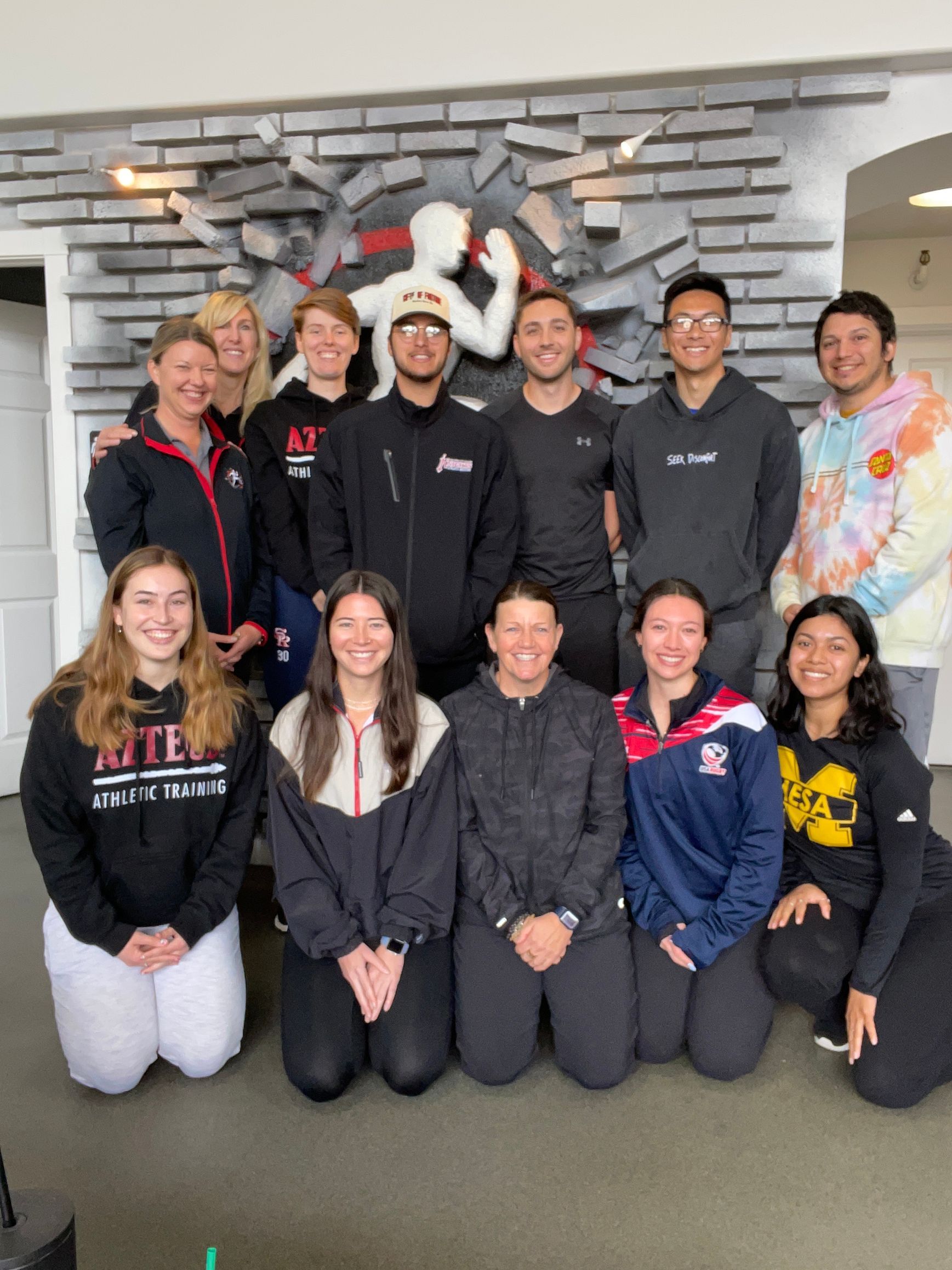 Group of people posing indoors in front of a stone wall, many wearing matching black sweatshirts.