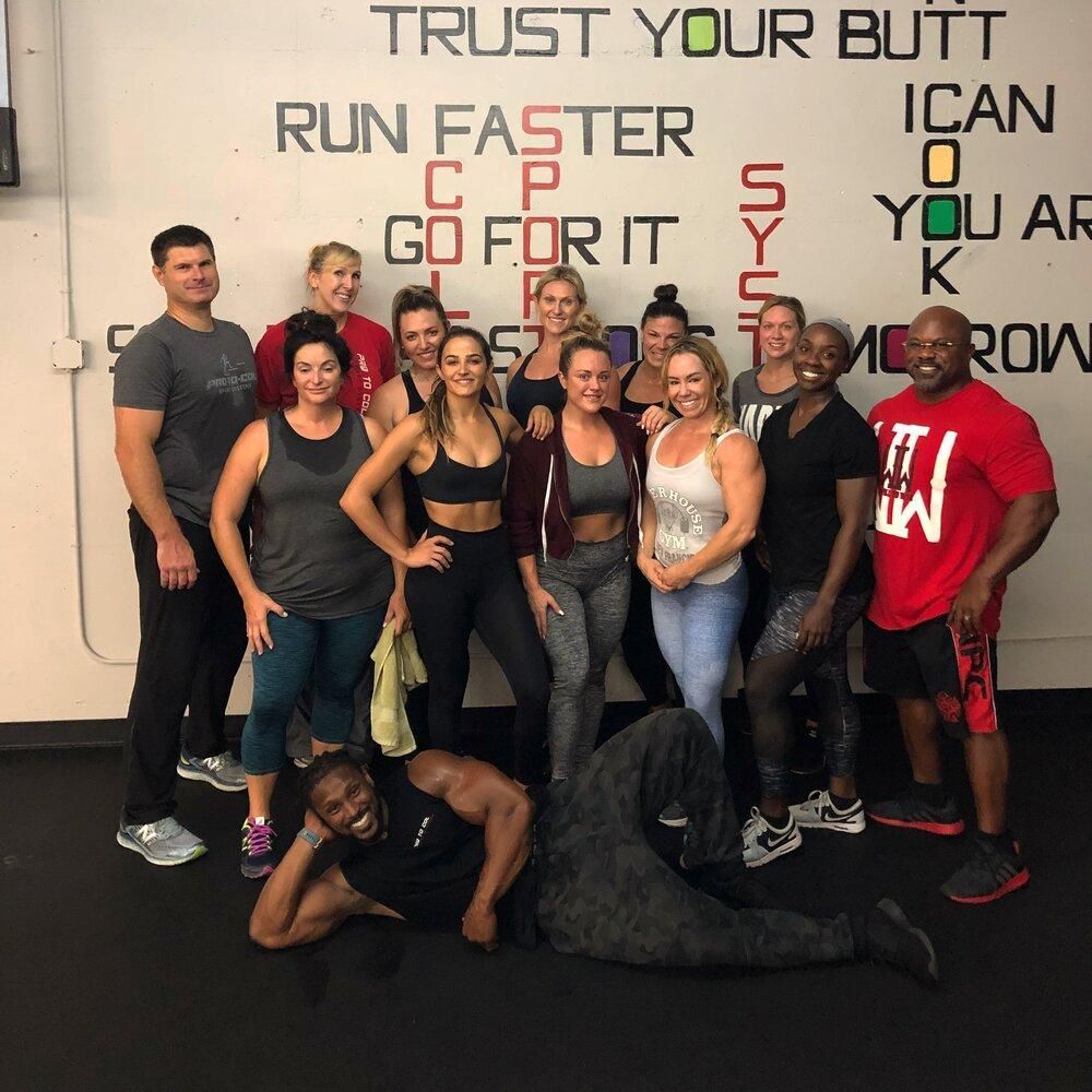 Gym group posing under motivational wall text, with one person lying on the floor in front.