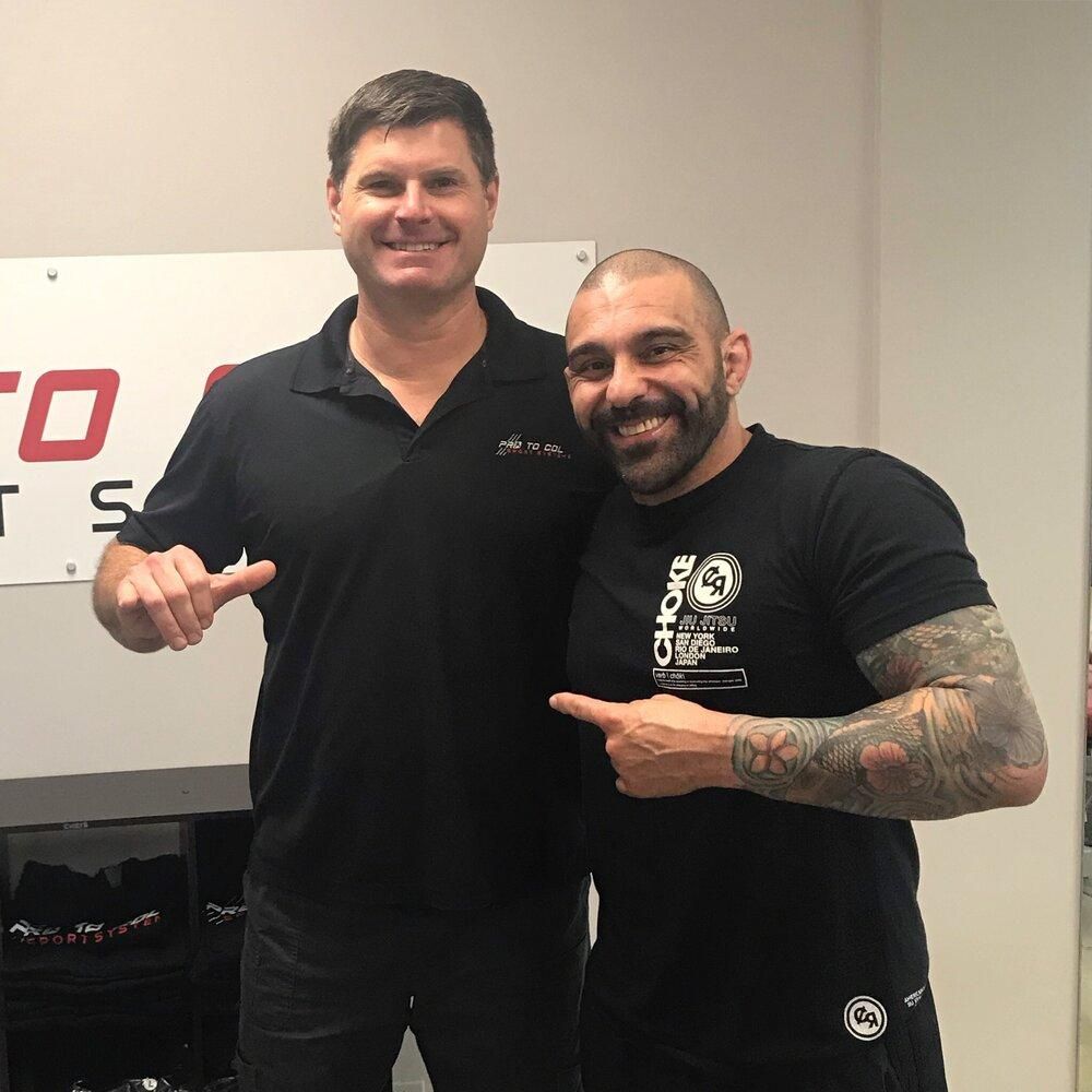 Two men smiling and pointing at each other indoors, one in a black polo and one in a black T-shirt.