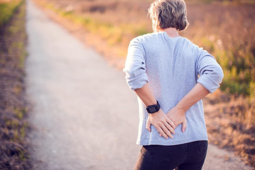 Person in a light blue shirt stretching on a path outdoors, holding their lower back