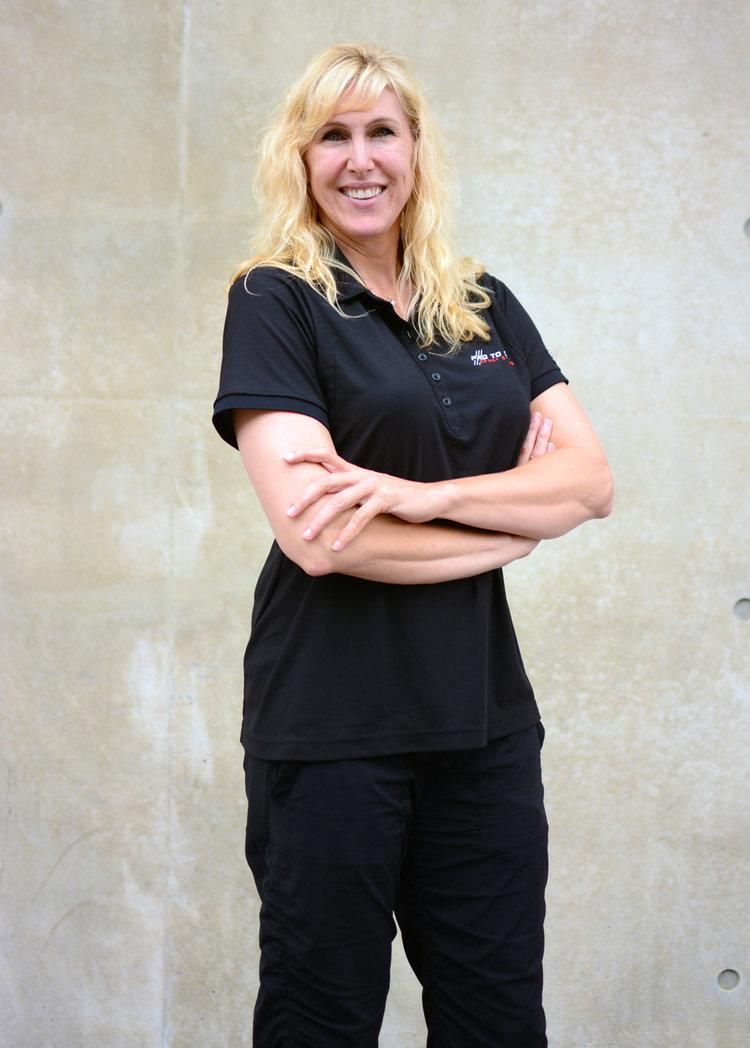 Smiling blonde person in black clothes standing with arms crossed against a beige wall