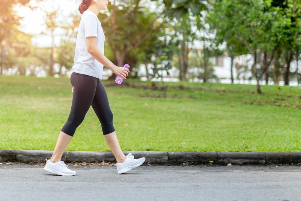Person walking on a sidewalk in a park, holding a purple water bottle, wearing black leggings and white sneakers.
