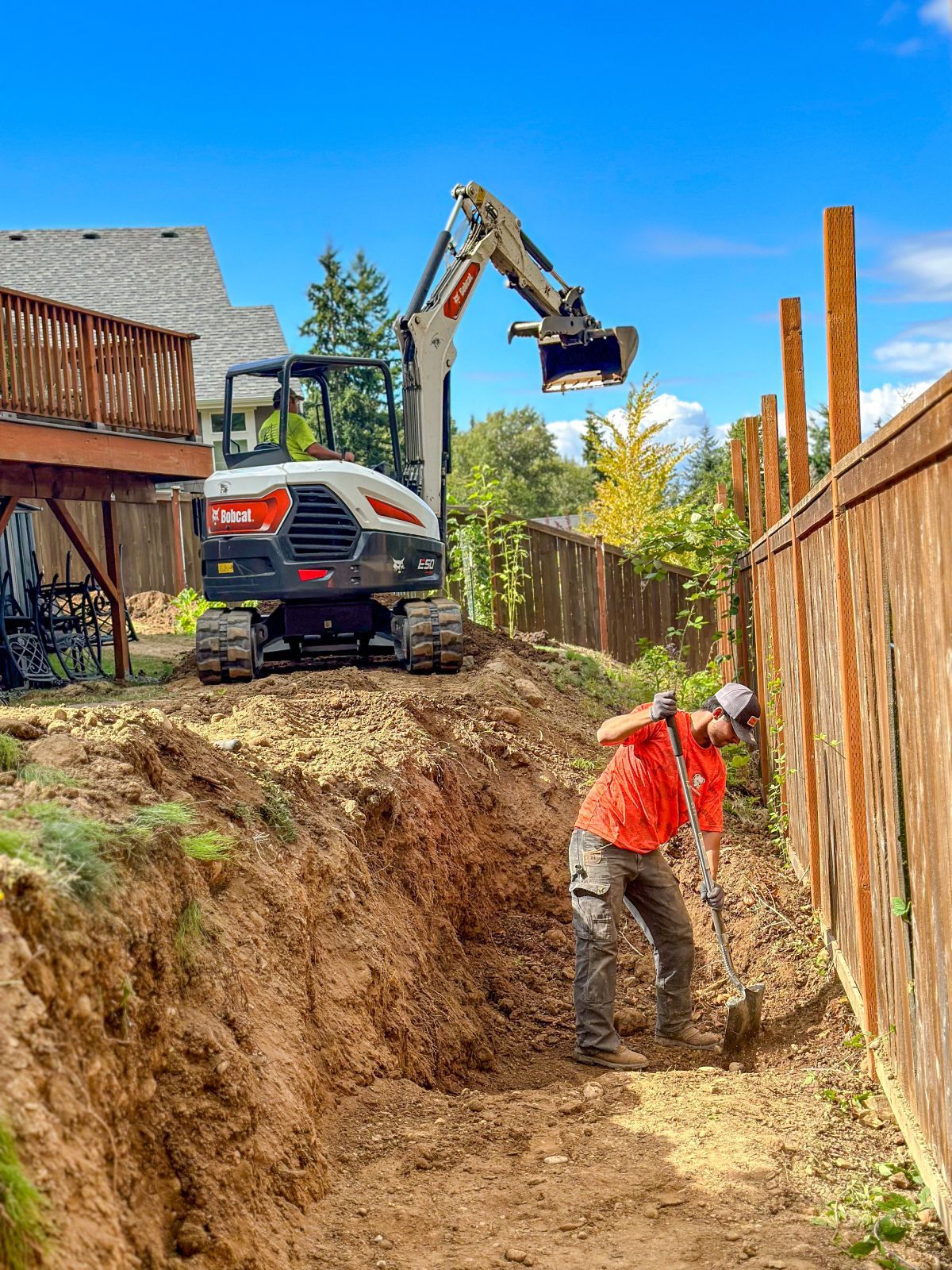 A worker digs near a wooden fence, while a Bobcat excavator digs in the background. Sunny day.