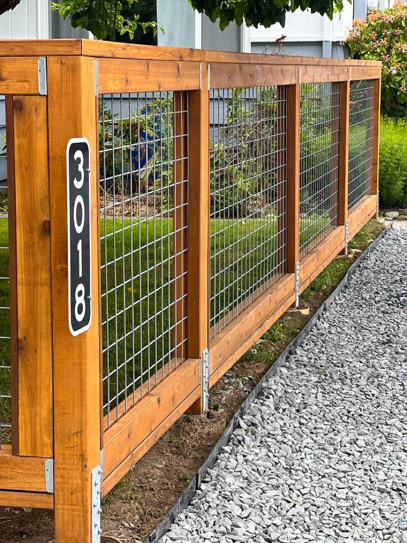 Wooden fence with metal wire panels, gravel path, and house number sign.