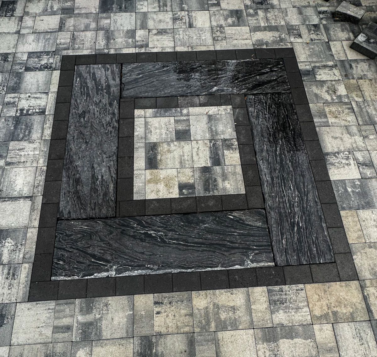 Square patio with dark granite stone inlay bordered by dark pavers on light pavers