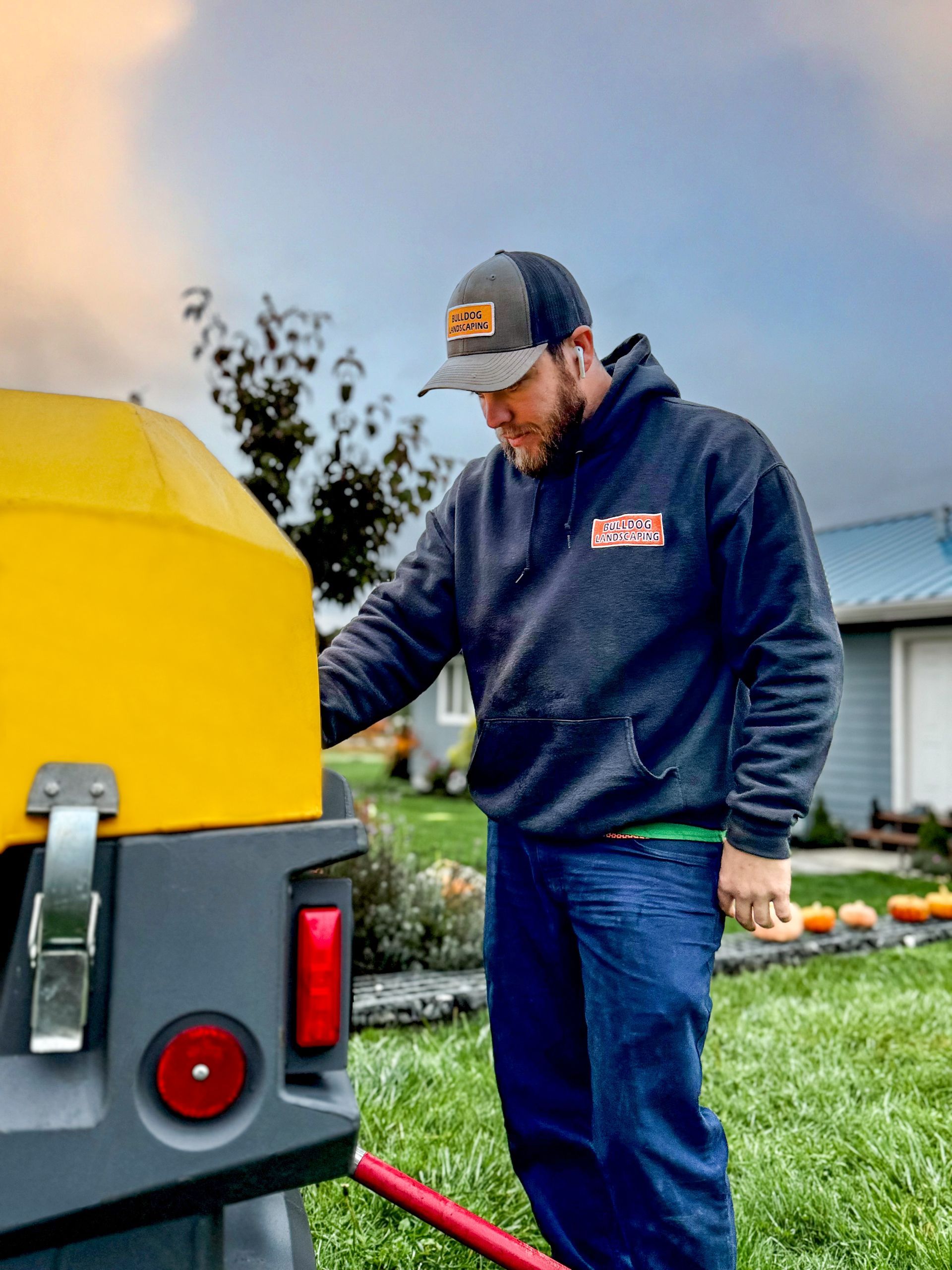 Man in a hoodie and cap working on a yellow trailer, with pumpkins and a house in the background.