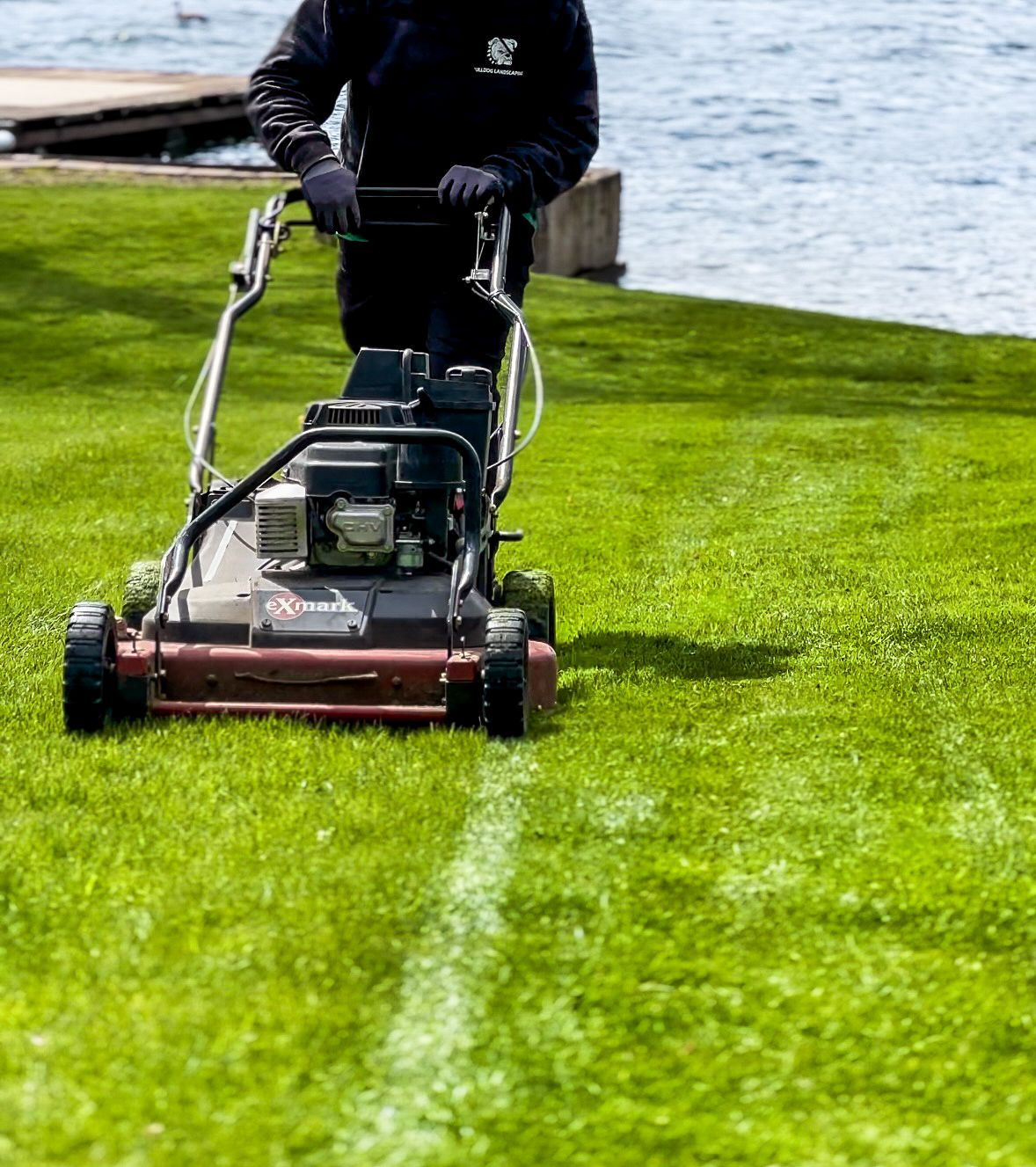 Person mowing lawn with a lawnmower near water, creating a defined stripe in the green grass.