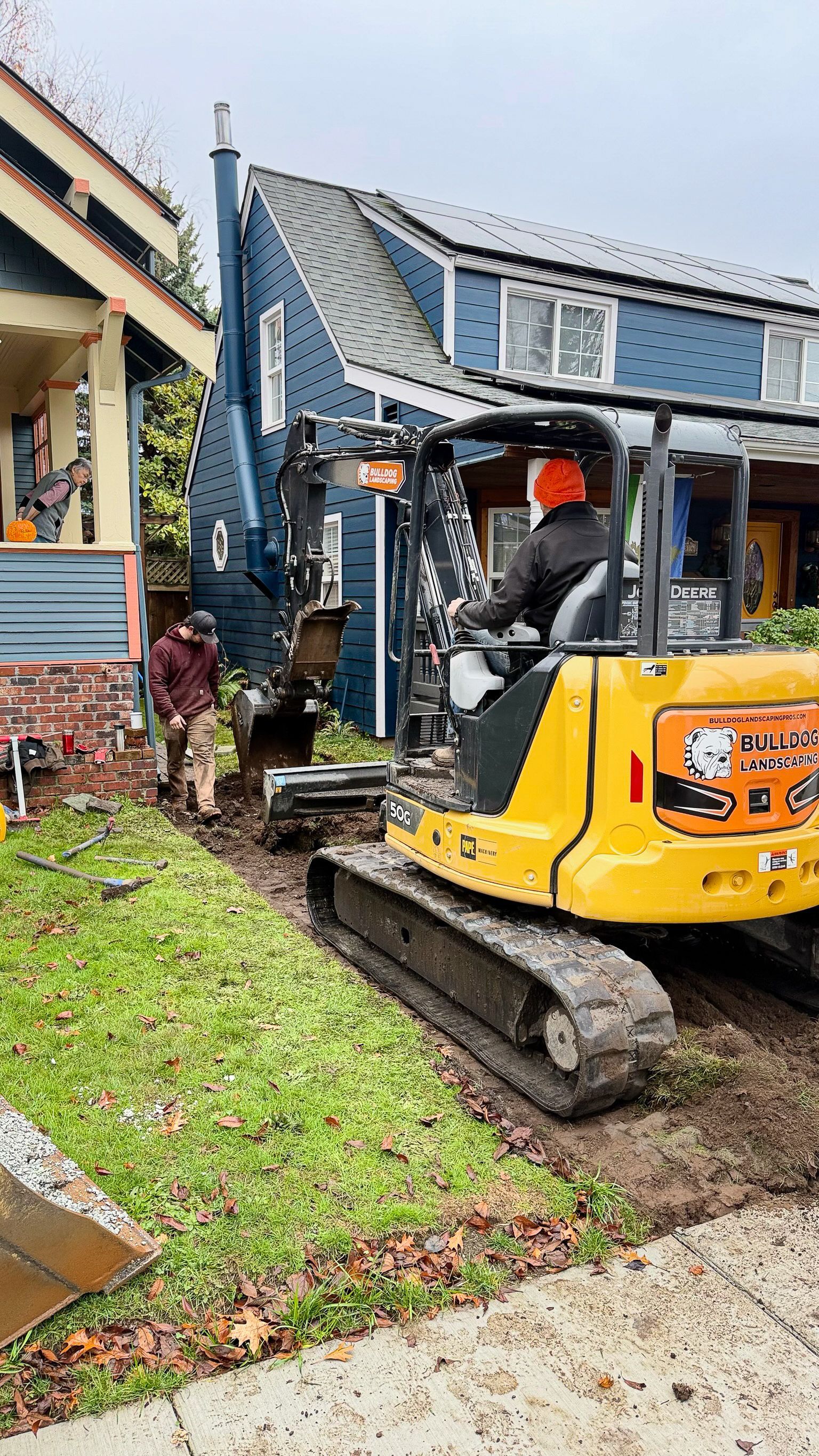 Mini excavator digging trench in front yard next to blue house; worker in orange hat operating it, another person standing by.