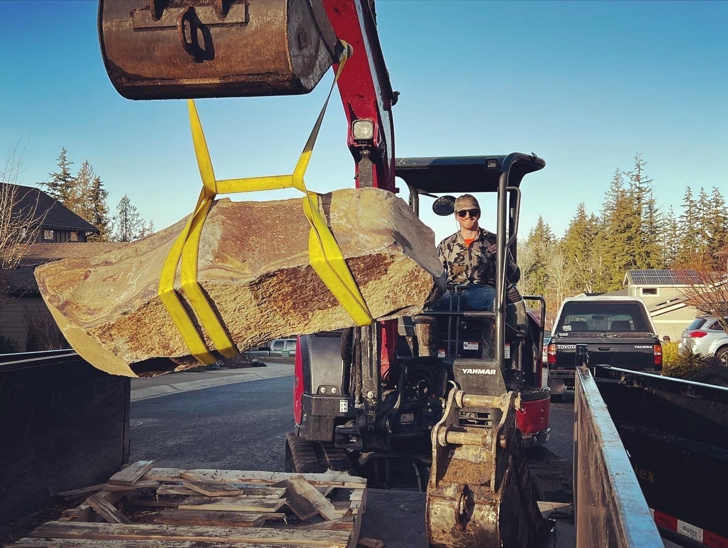 Tractor lifting a large rock slab with yellow straps to load into a dump truck. Person in driver's seat.