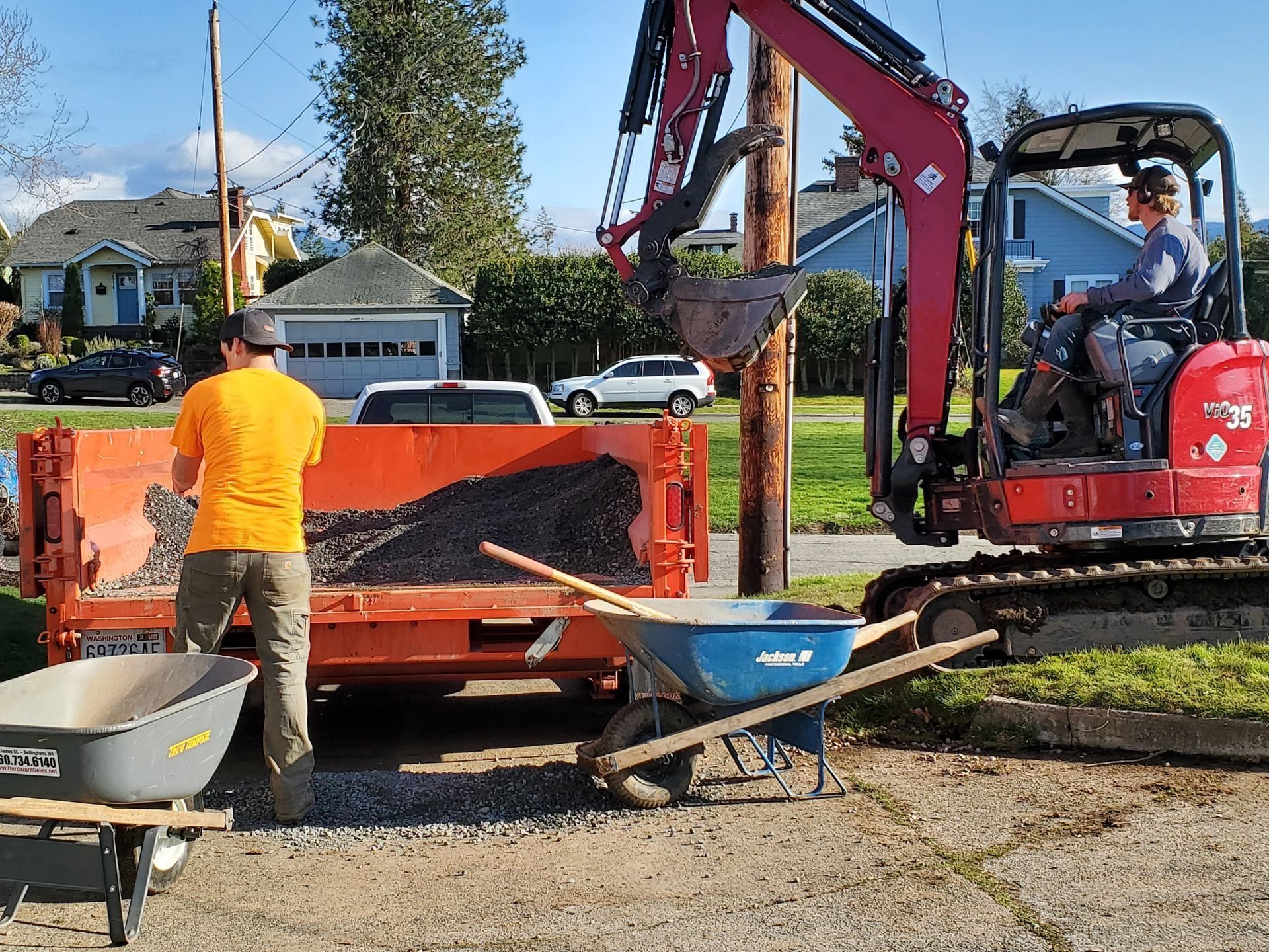 Construction workers using an excavator and dump truck to move gravel. One worker standing, another operating the machinery.