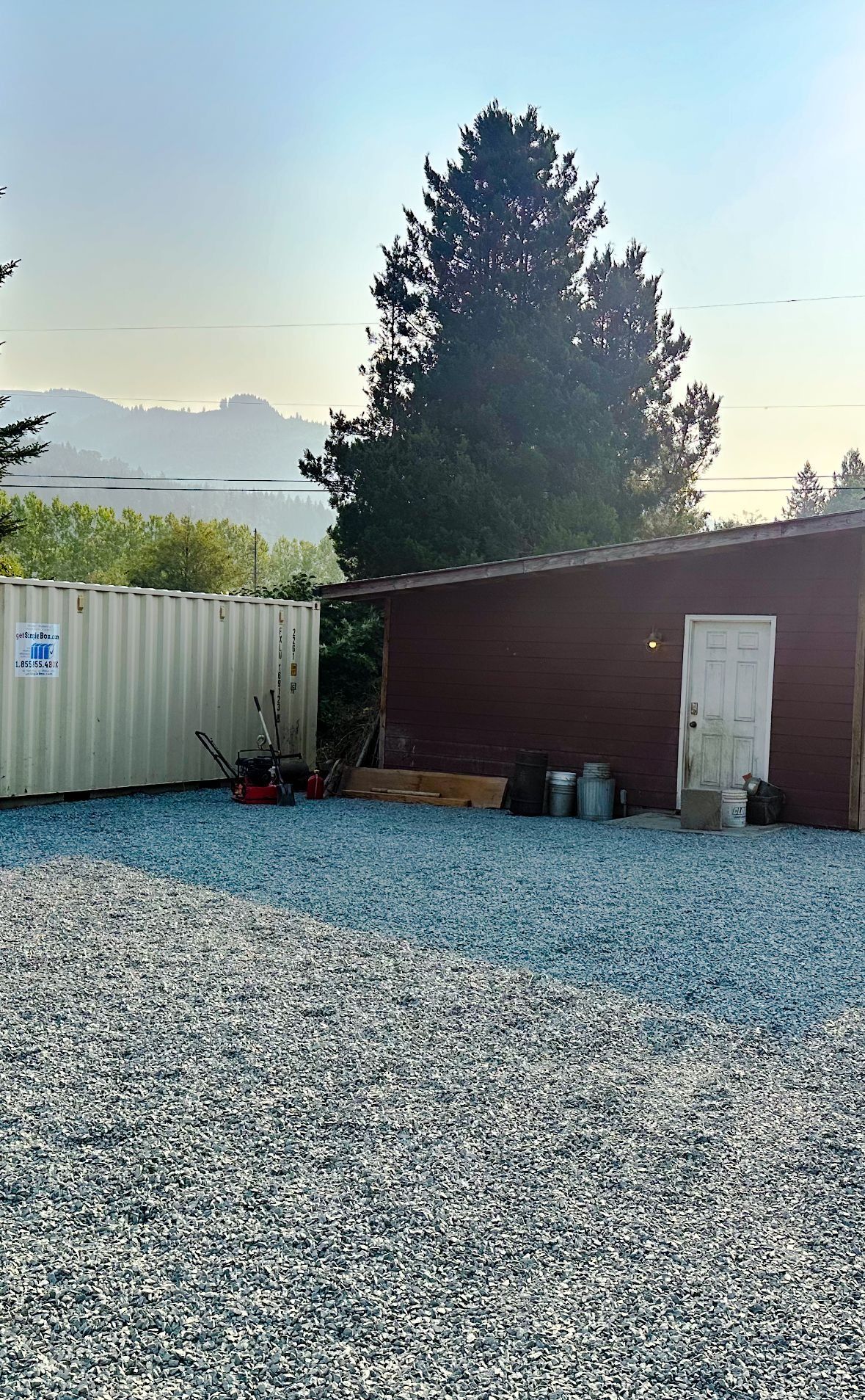 A gravel yard with a shed and storage container; mountains and a tall tree in the background.