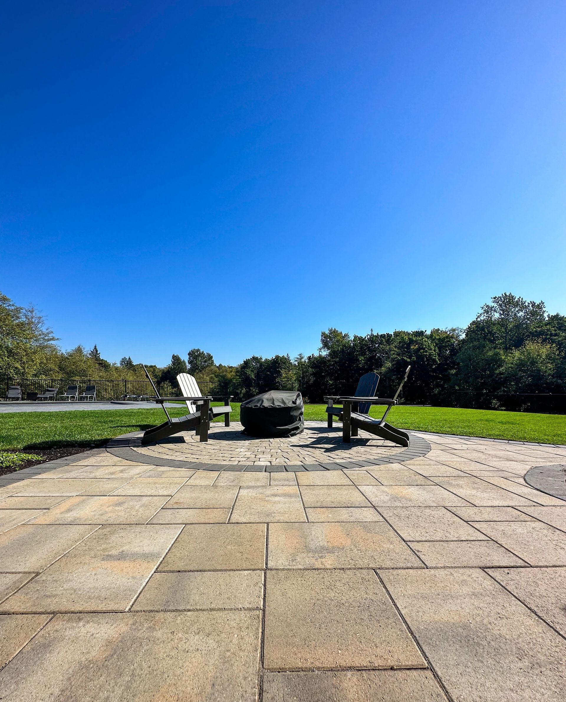 Two chairs face a fire pit on a stone patio under a bright blue sky. Green grass and trees are in the background.