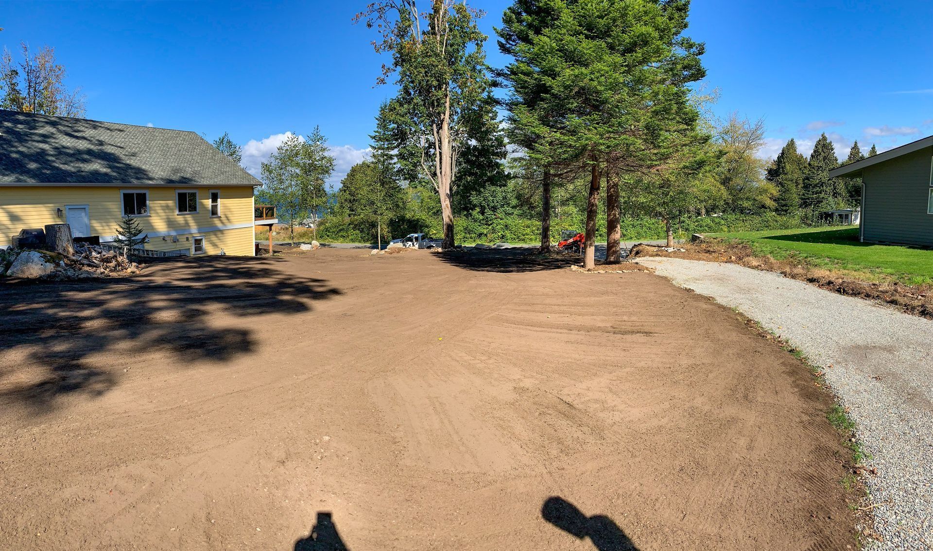 A cleared, dirt yard next to gravel pathway and yellow house, under a blue sky with trees.