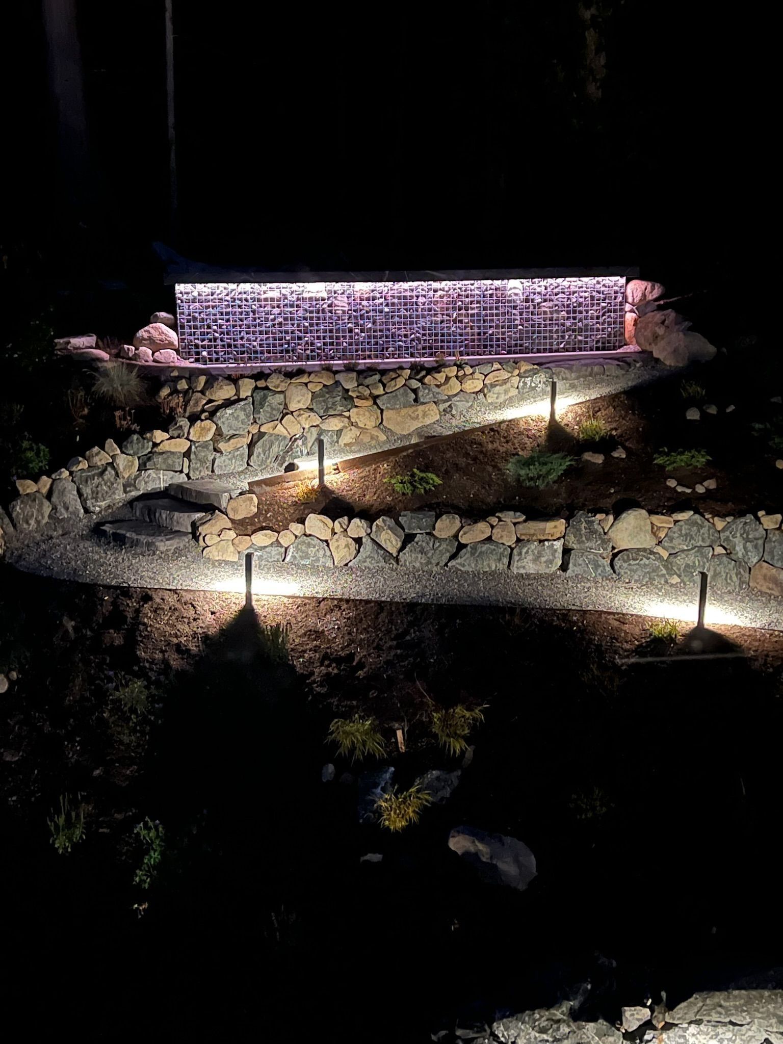Lit-up stone structure at night on a hillside. Pathway with lights leads up. Dark surrounding.
