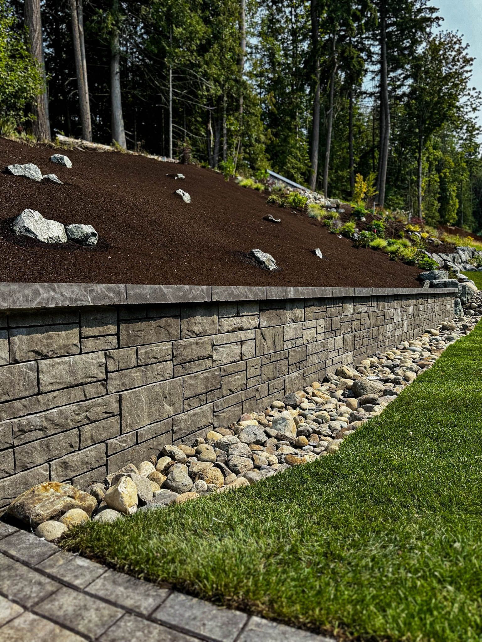Stone retaining wall with a grassy lawn, rocks, and a mulched hillside.