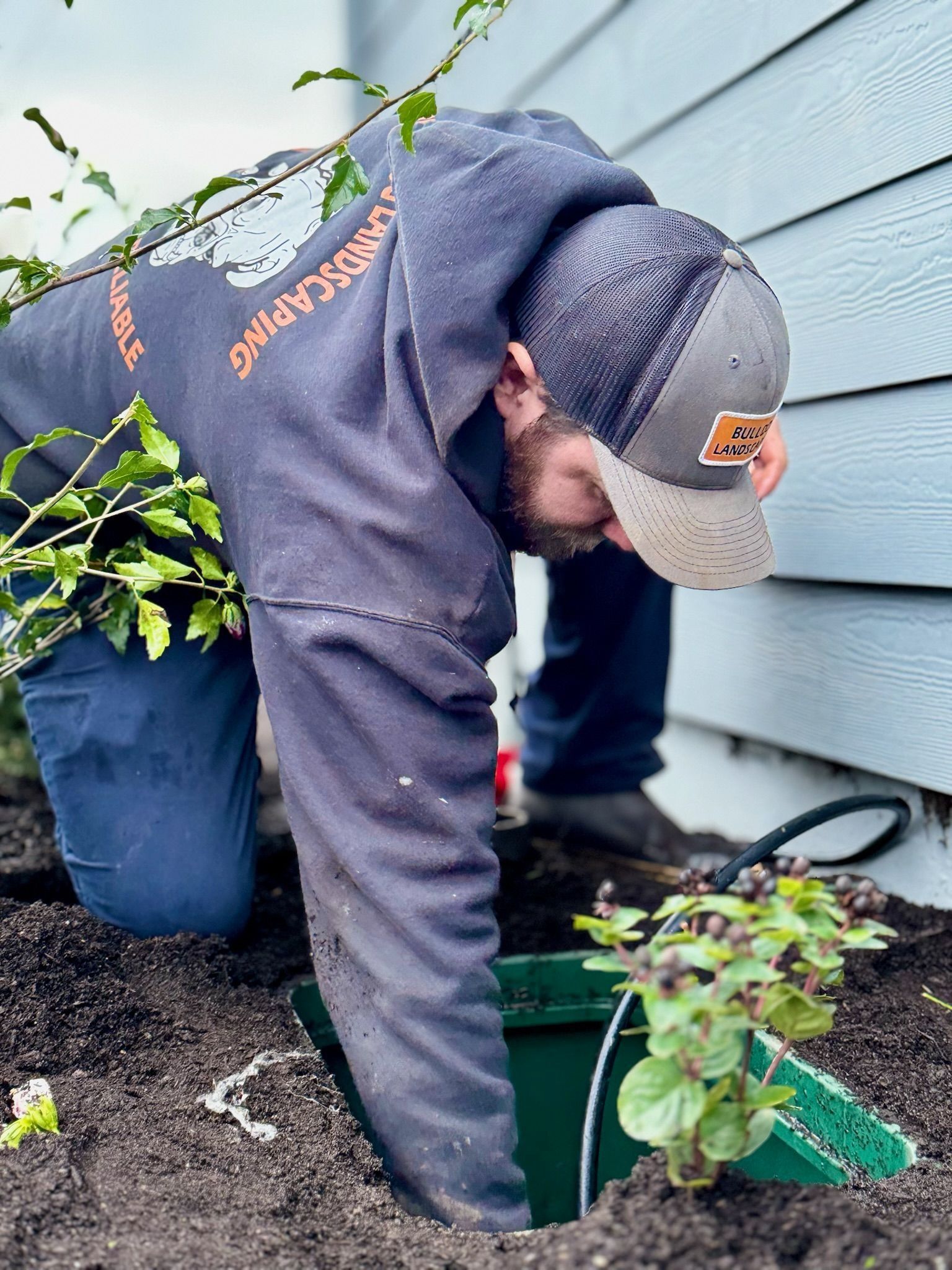 Man kneeling, planting in a garden near a building. He wears a hat and hoodie, holding a small plant and bucket.