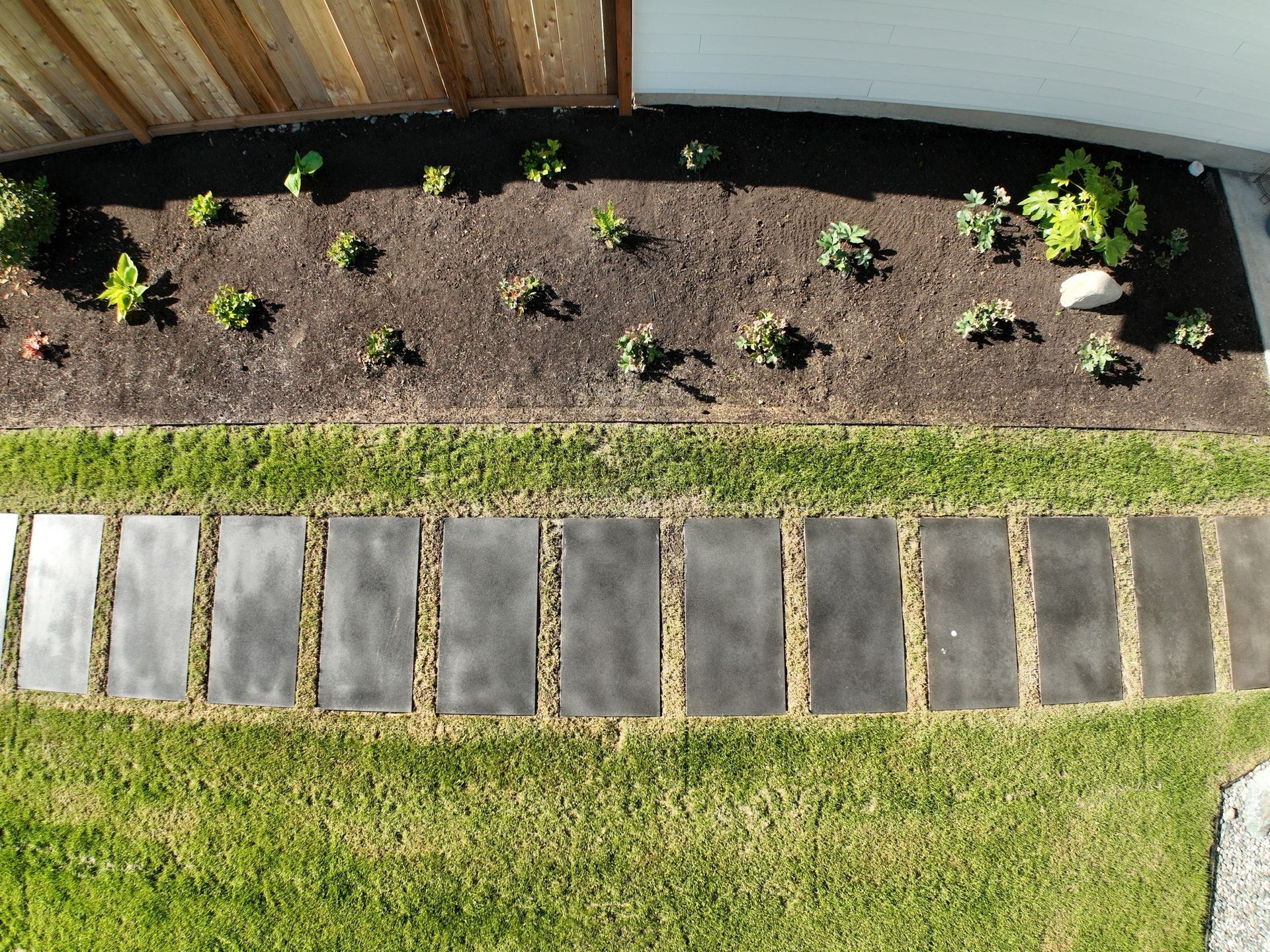 Path made of gray rectangular stones in a green lawn, bordering a garden bed with young plants and dark soil.