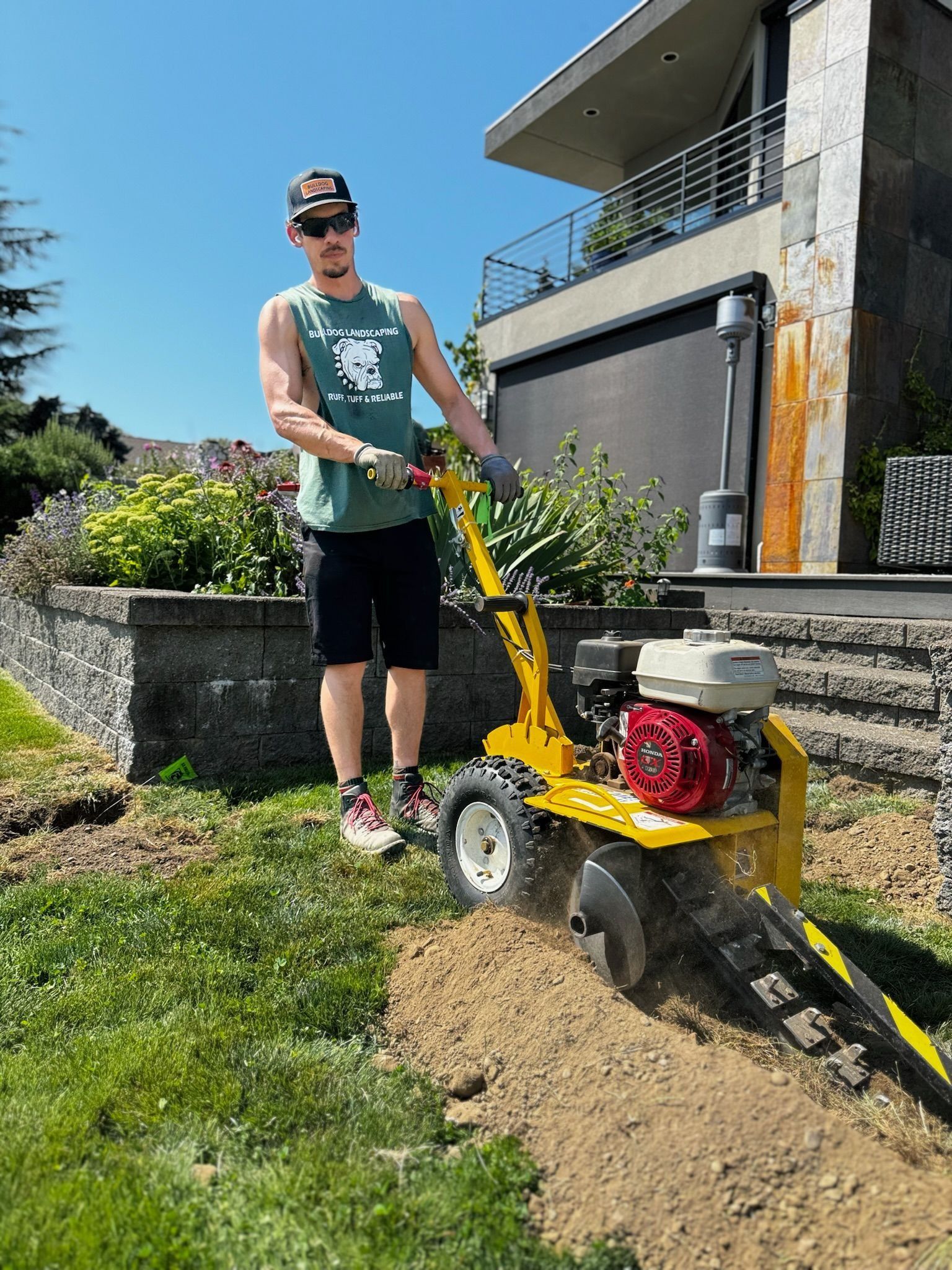Man operating a yellow trenching machine, digging a trench in a grassy yard, under a sunny sky.