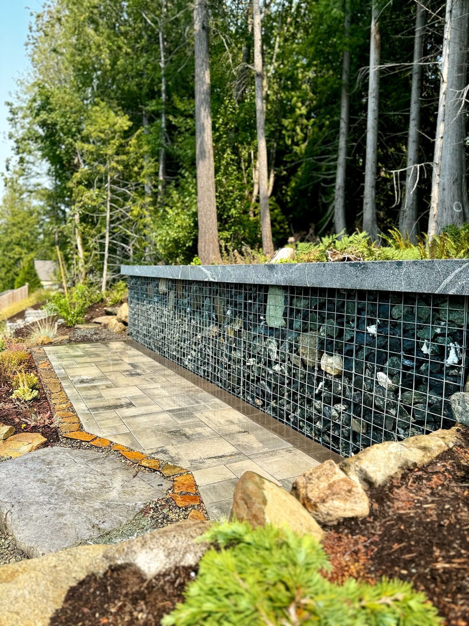 Stone path leading to a retaining wall filled with dark rocks, surrounded by trees and greenery.