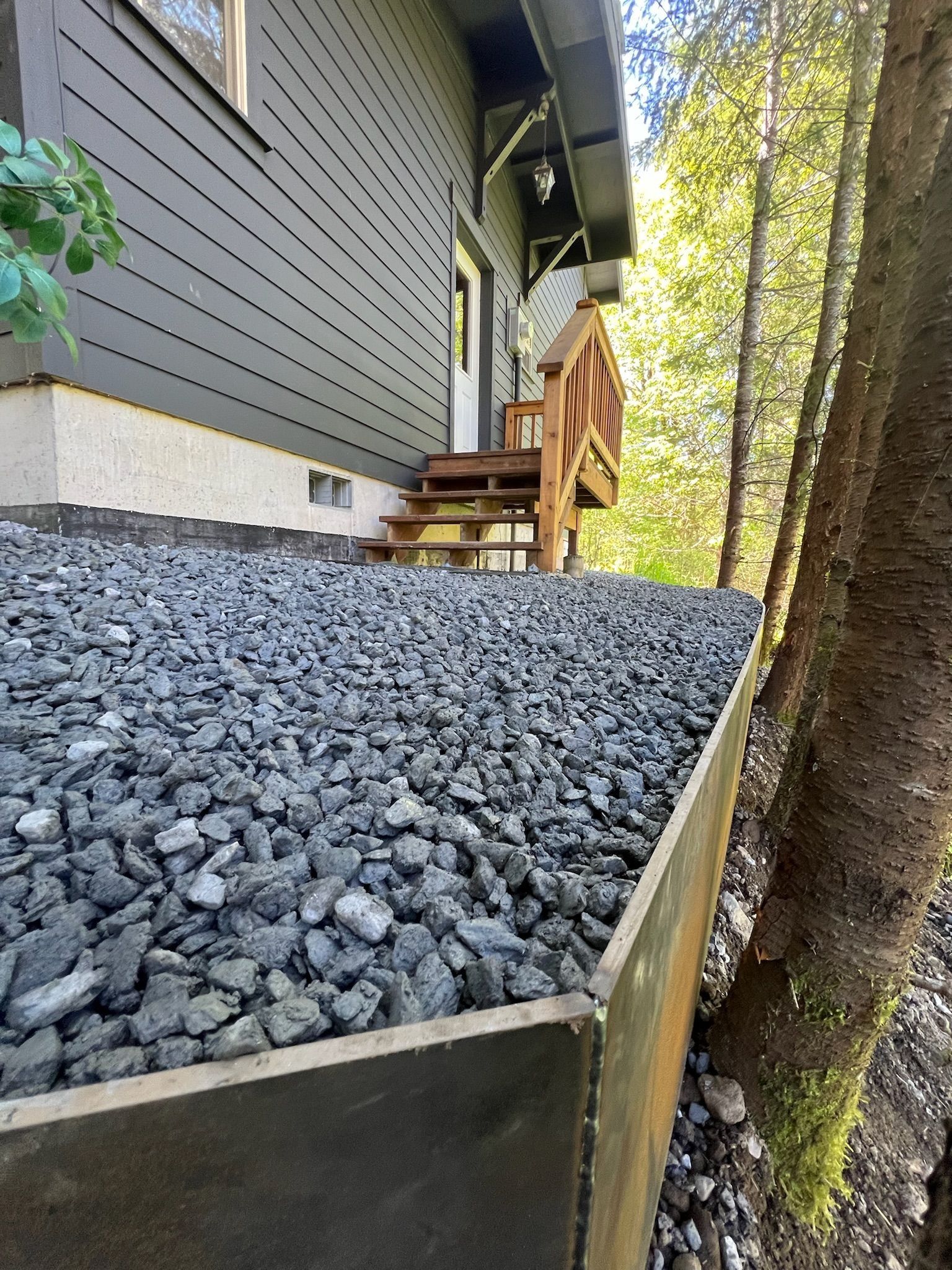 Gravel-covered slope alongside a gray house with wooden stairs and railing. A tree stands on the right.