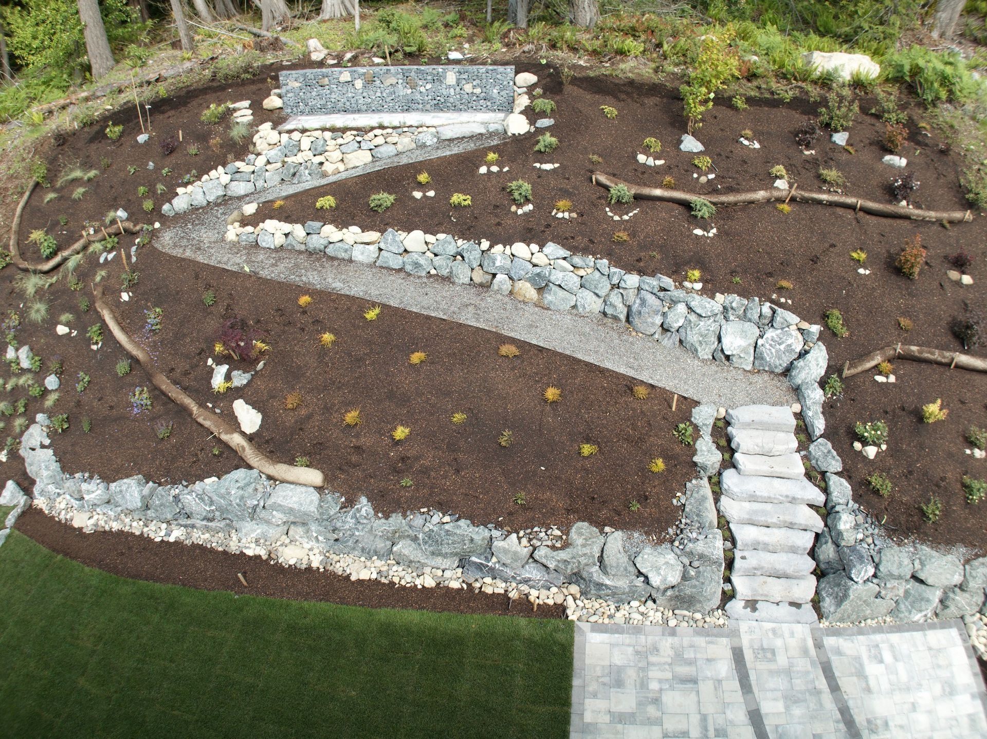 Elevated garden with a winding rock path, stone steps, mulch, and greenery; a small water feature is at the top.