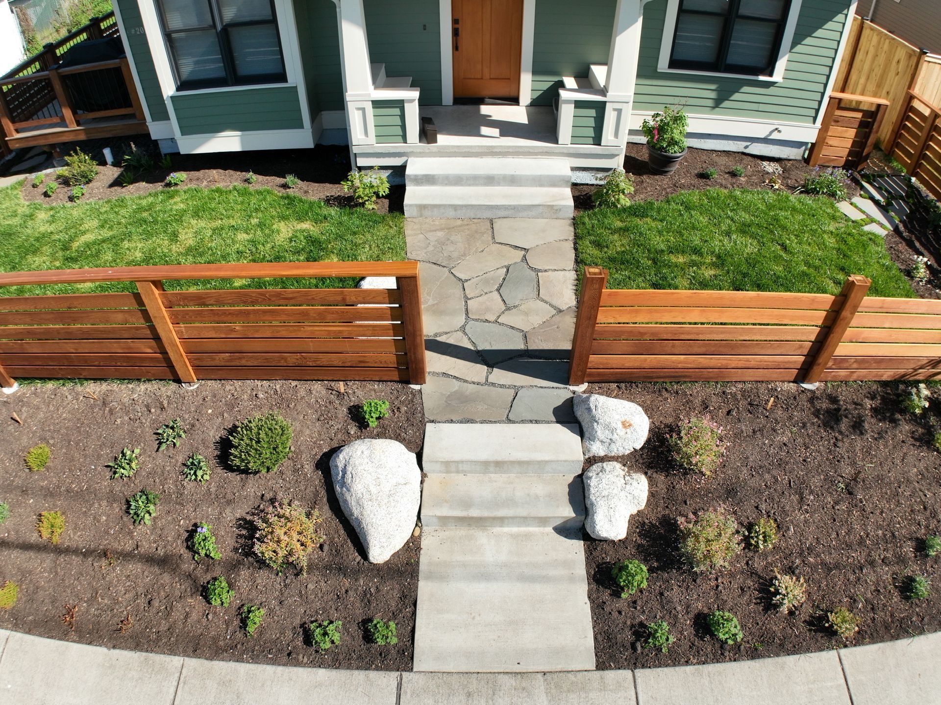 Front yard with stone path leading to a green house, flanked by wooden fences and planted beds.