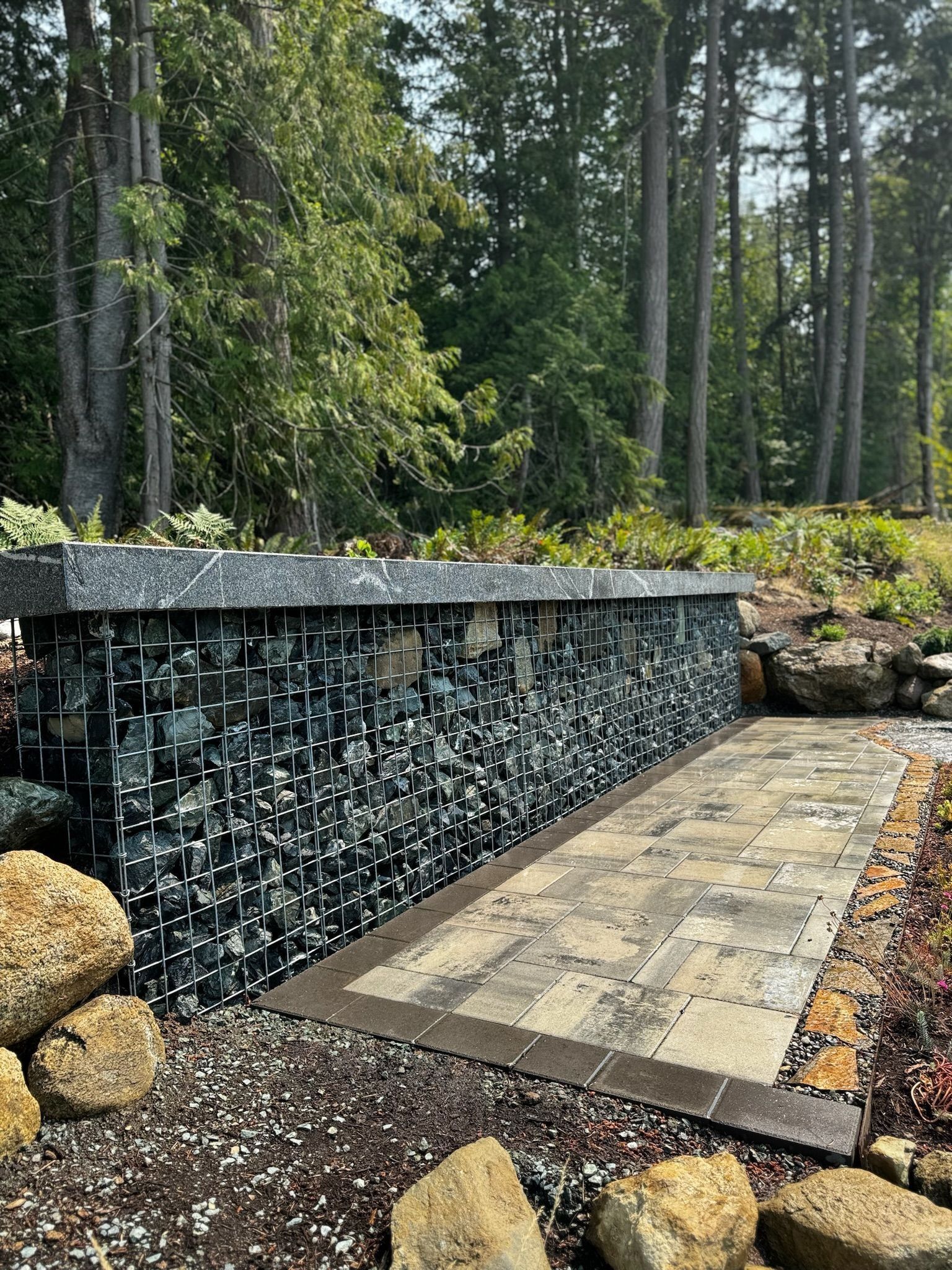 Gabion wall filled with dark rocks, bordering a stone pathway in a forest setting.