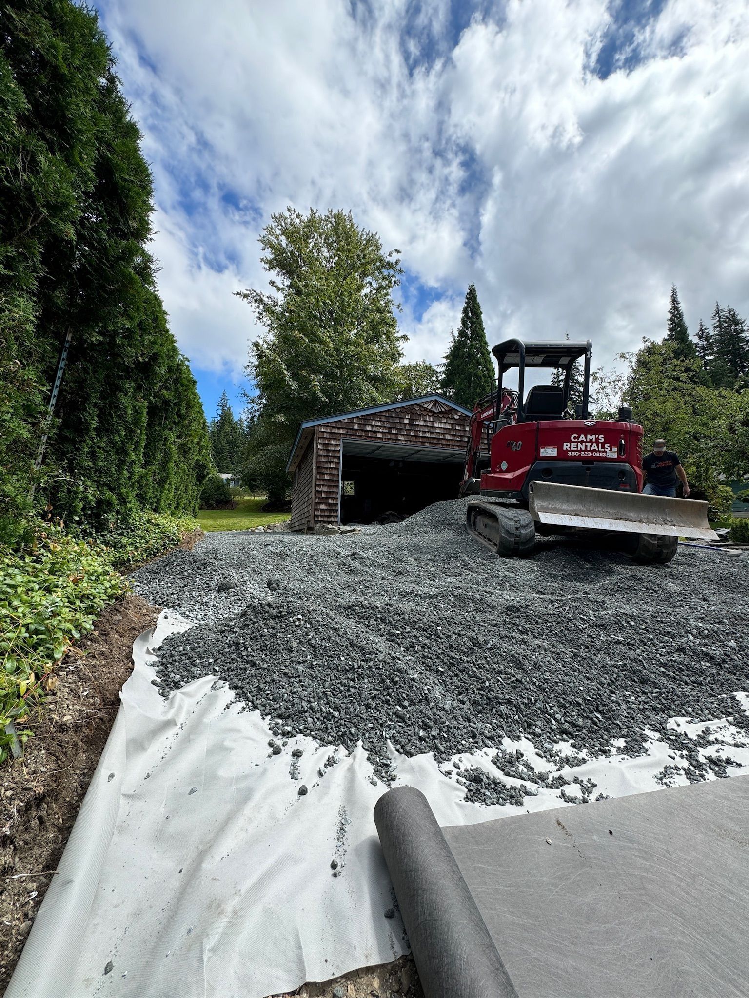 Gravel being spread by a red tractor on a driveway, with a shed in the background and cloudy sky above.