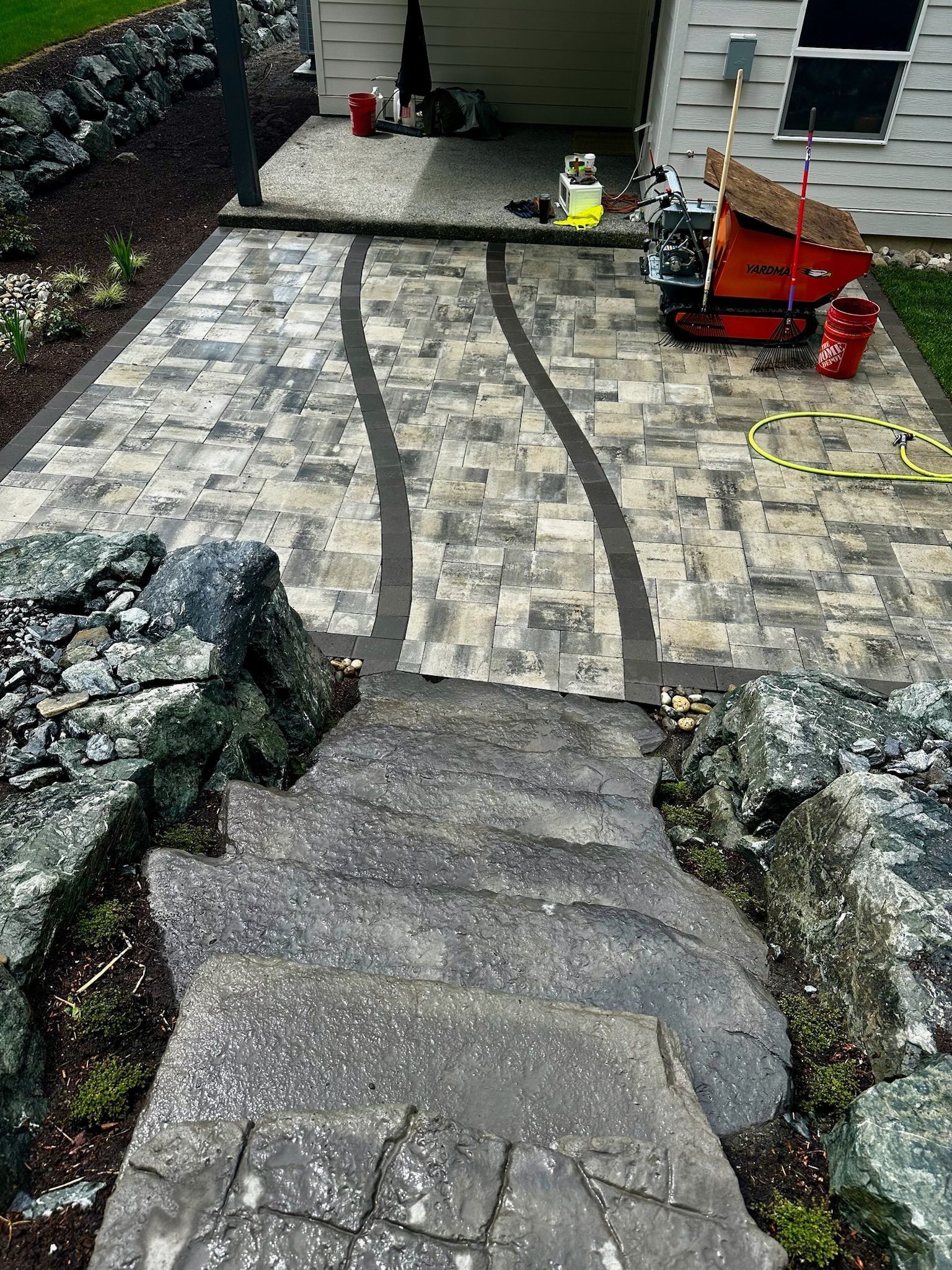 Stone steps lead to a paved patio with winding dark border. Equipment and shed in the background.