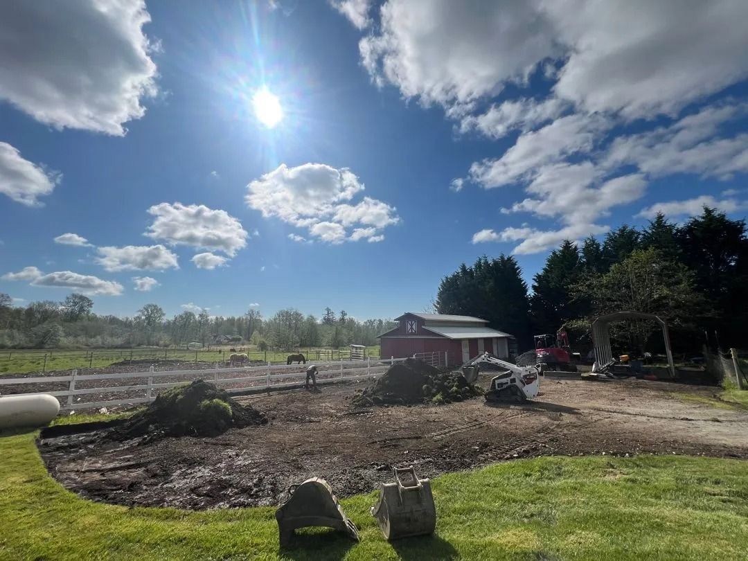 Bright sunny day over a rural property. Barn, trees, and dirt patch visible.