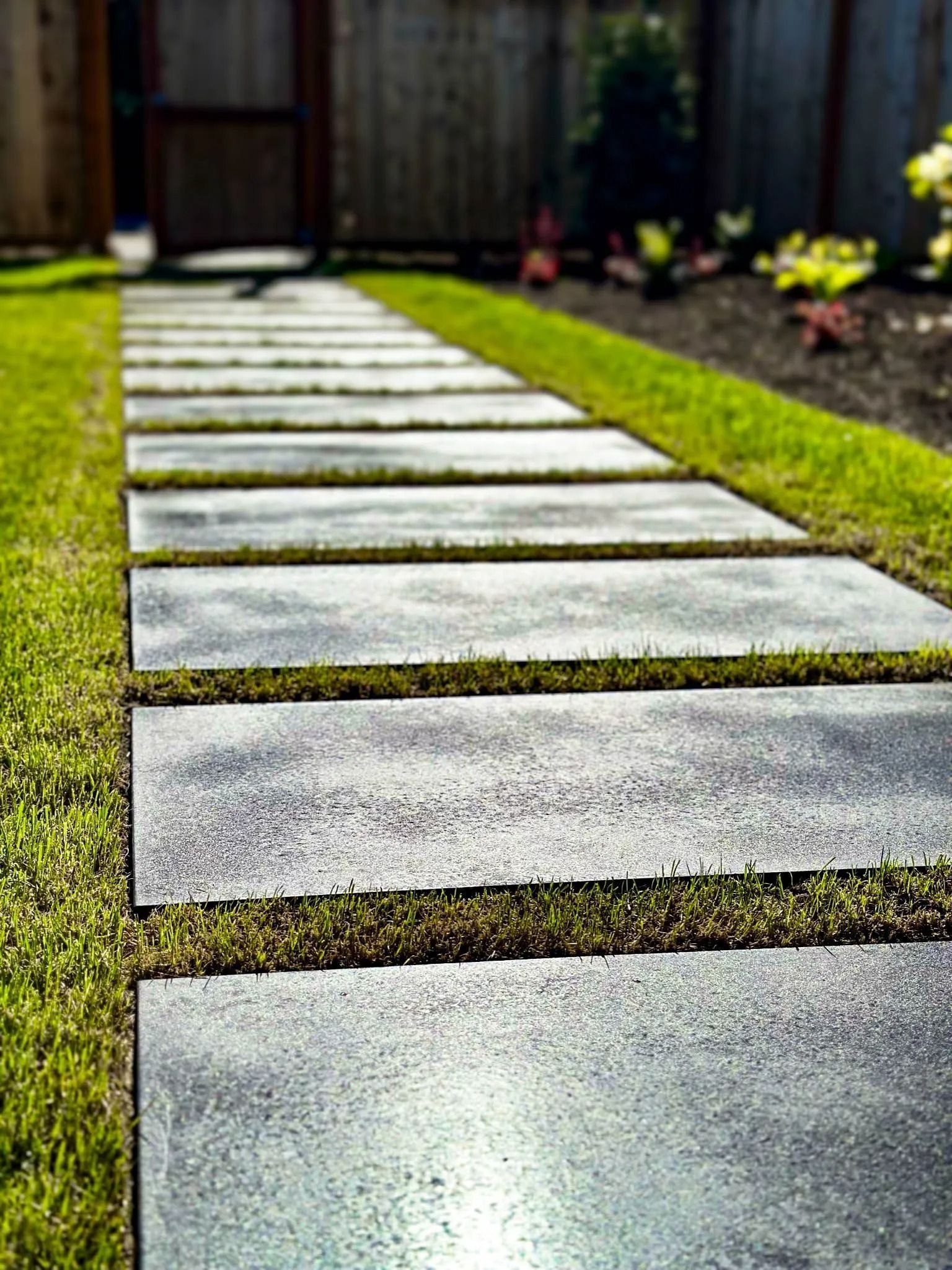 Stone pavers form a path through green grass, leading towards a wooden fence.