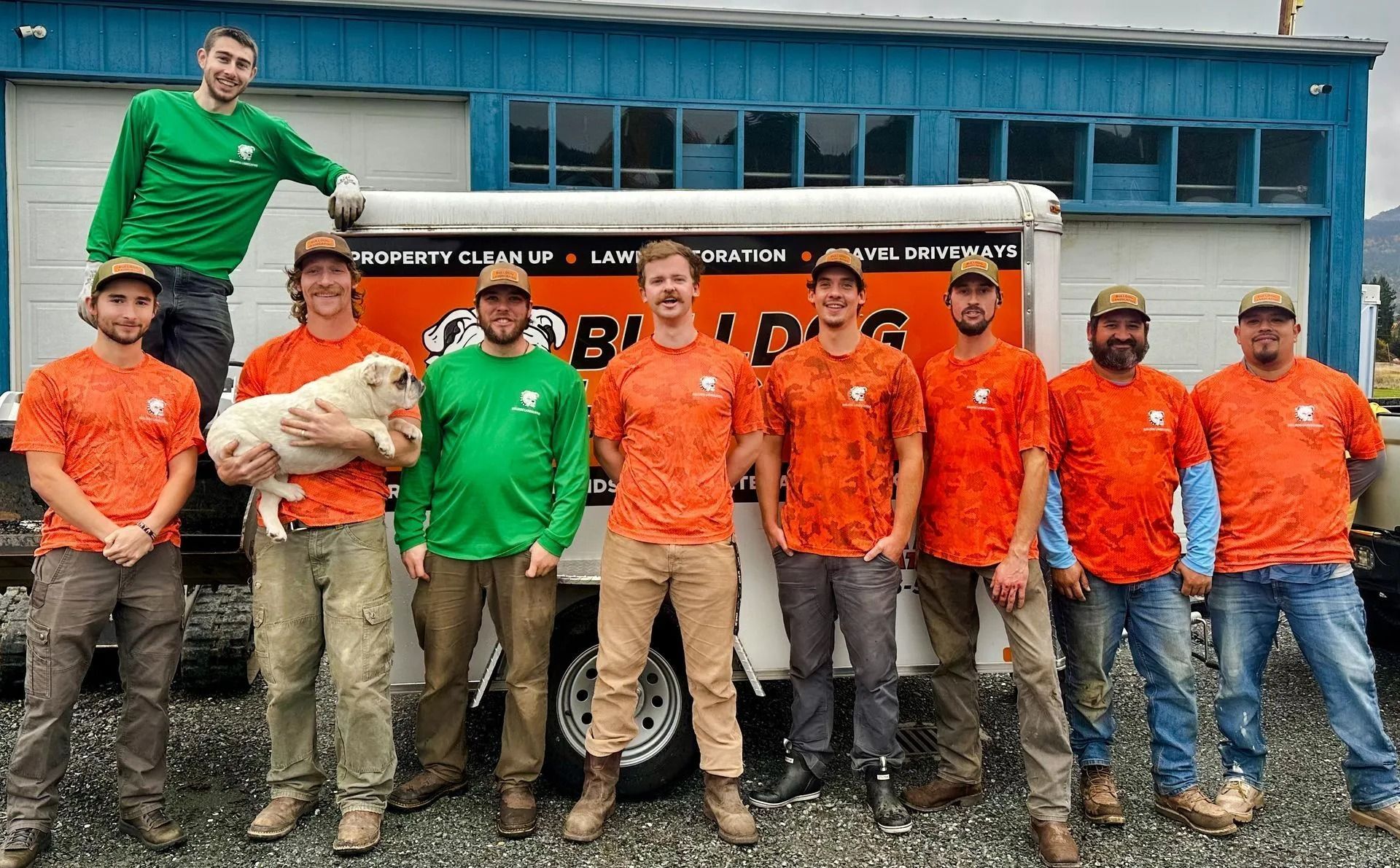 Construction crew poses in front of their company truck. Men in orange and green shirts; one holds a small dog.