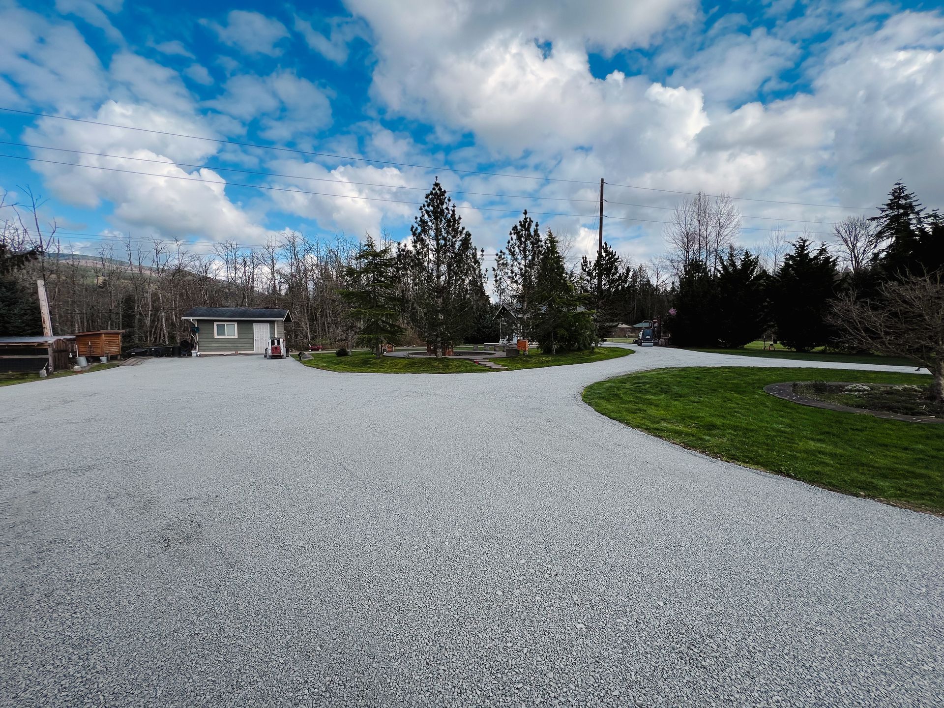 Gravel driveway curves to small building and trees, under cloudy blue sky