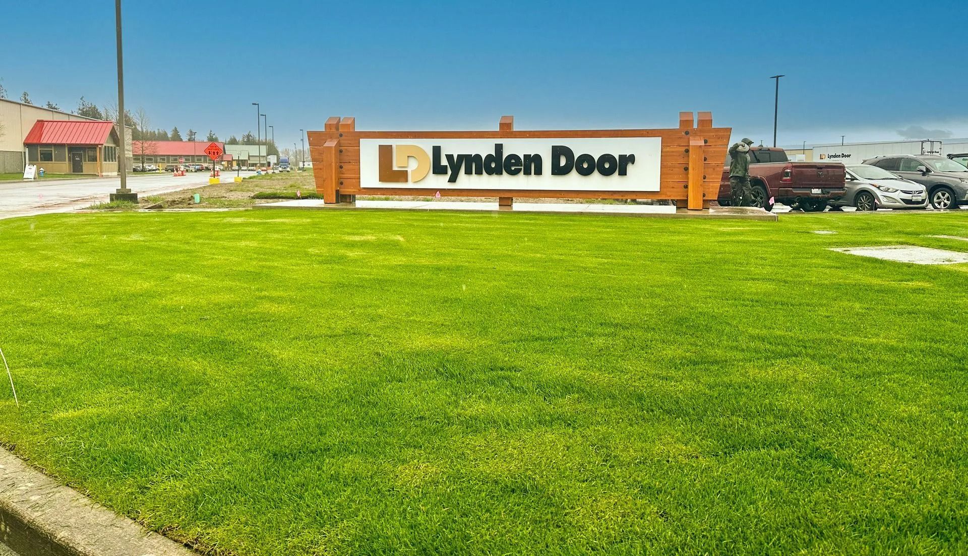 Lynden Door sign in front of green grass, with buildings and vehicles visible in the background on a sunny day.