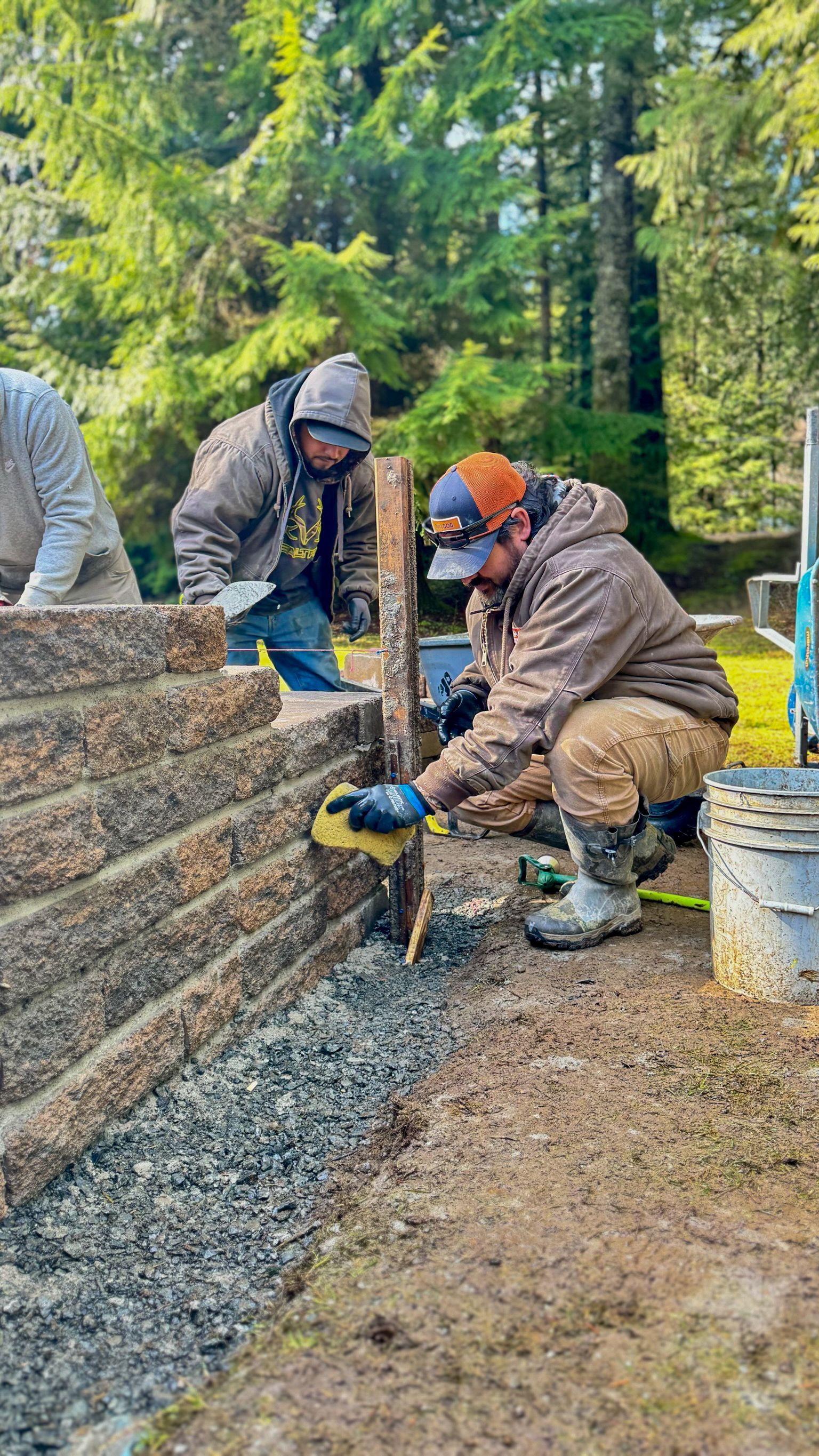 Workers using a jackhammer beside a stone wall at a wooded construction site