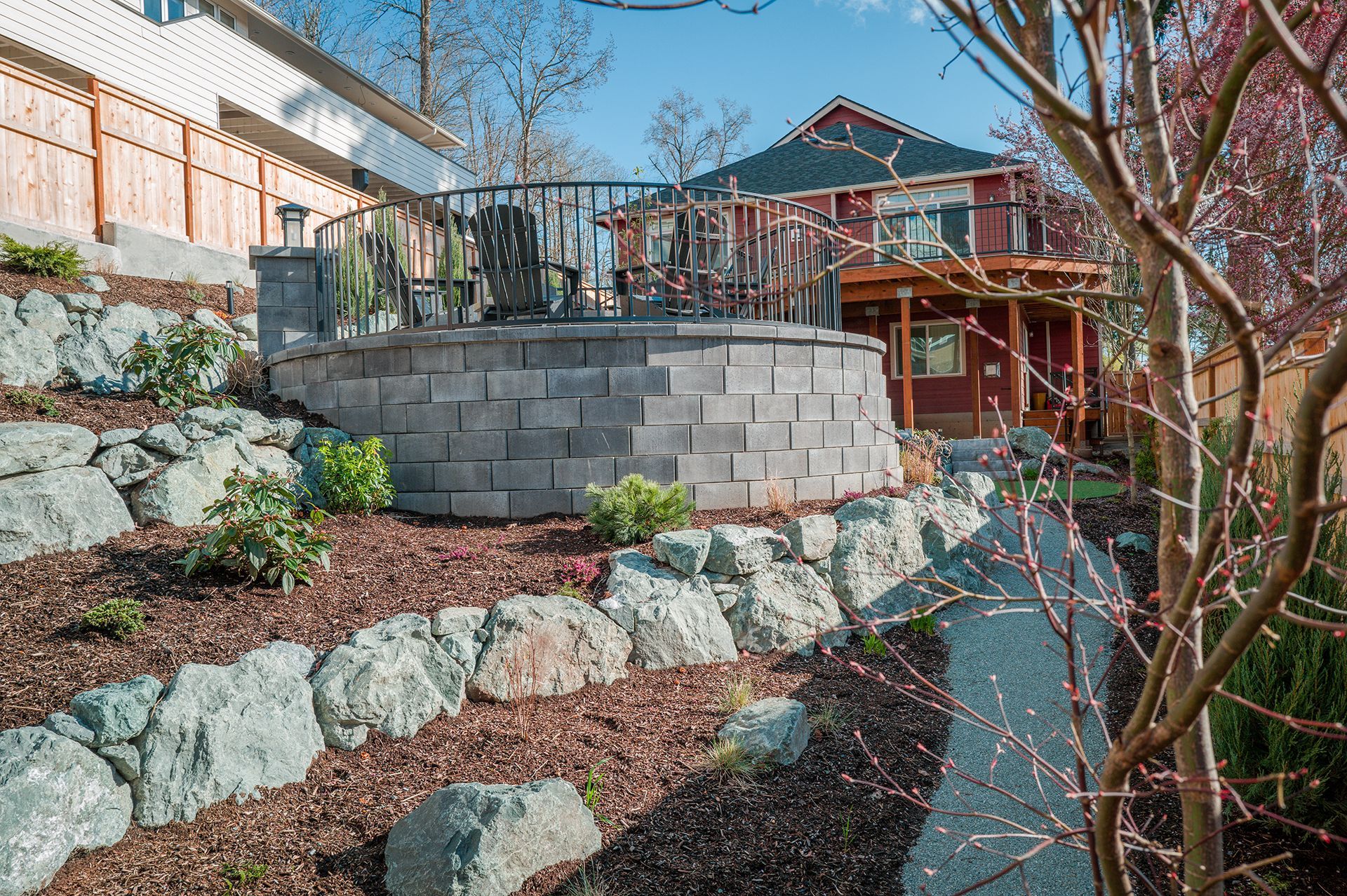 Terraced stone garden with retaining walls, shrubs, and a wooden deck beside a house.