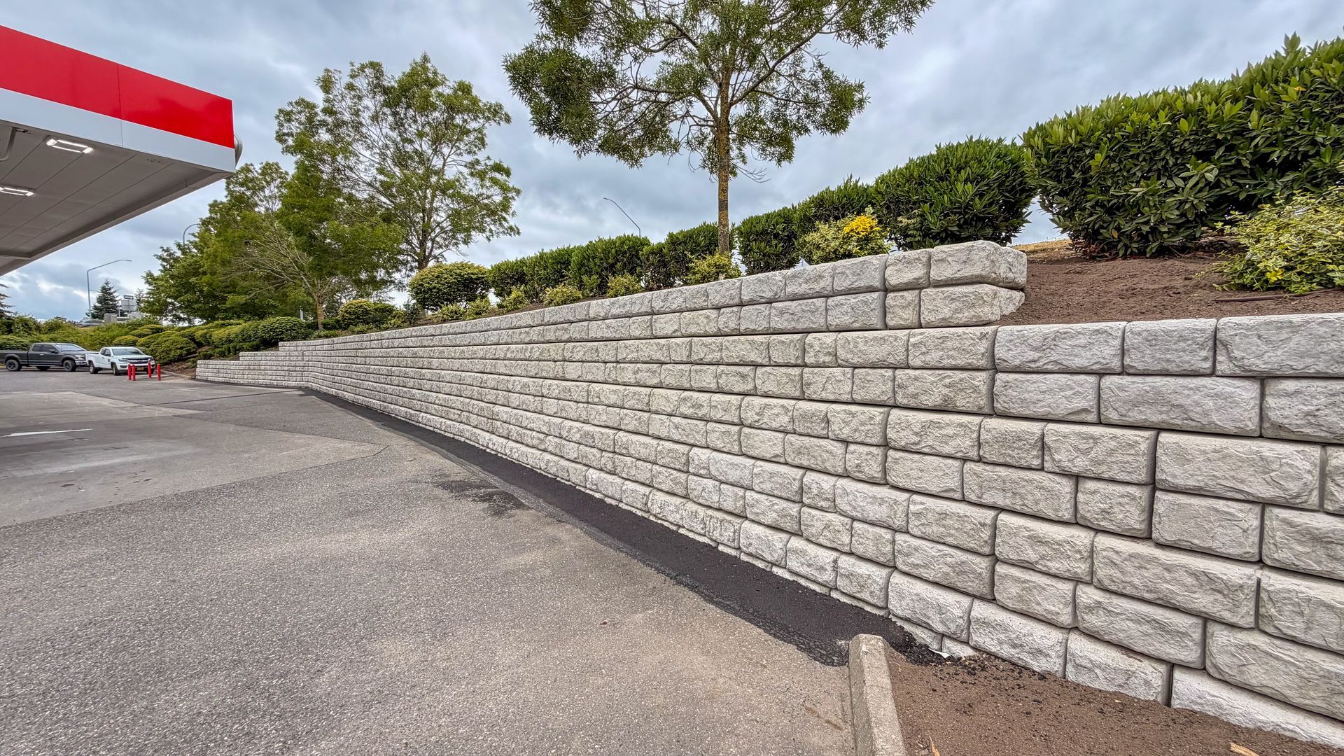 Empty parking lot beside a tall gray stone retaining wall with shrubs and trees above it.