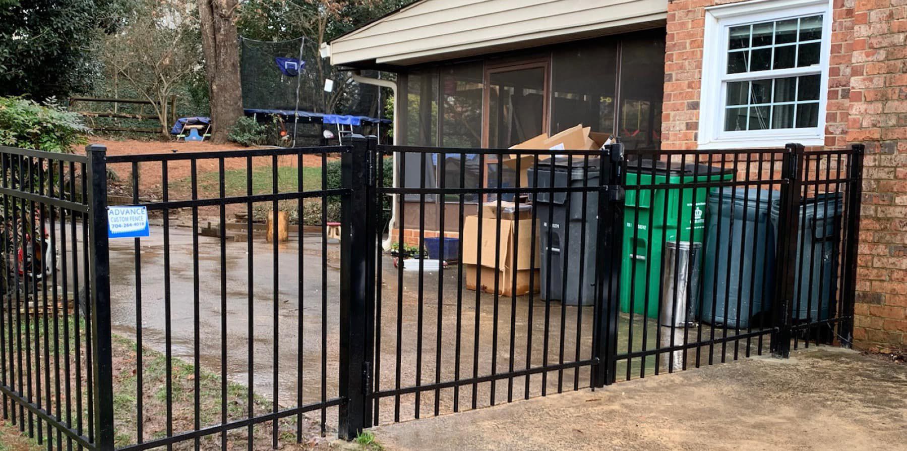 A black fence is surrounding a brick house with trash cans in front of it.