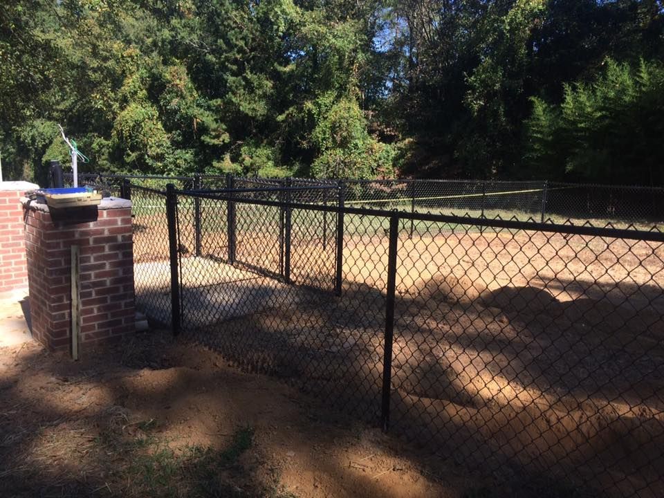 A chain link fence surrounds a dirt field with trees in the background.
