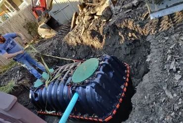 Man and excavator lower a large black septic tank into a trench.
