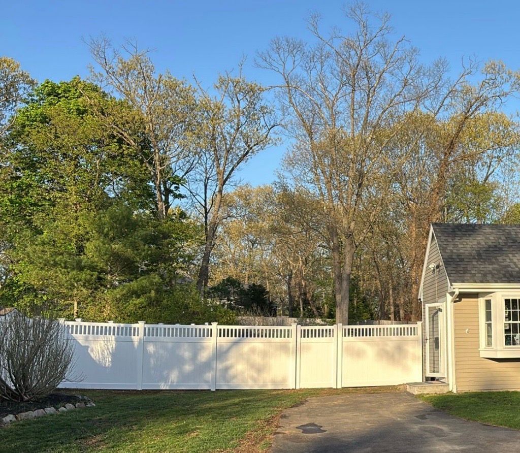 White fence along a driveway, trees in the background, and a section of a beige house on the right.