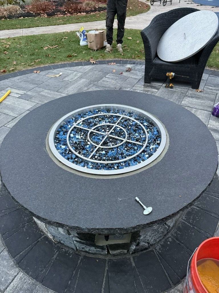 A circular stone fire pit sits on a patio, filled with blue glass fire beads, with a metal key resting on the top edge.