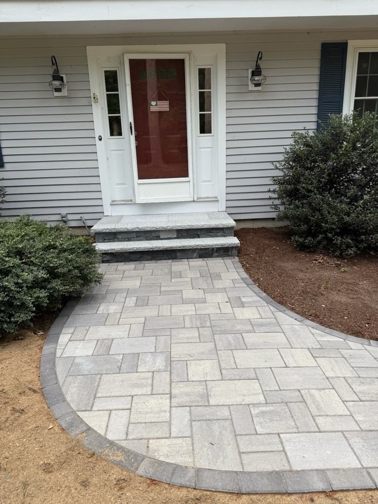 A light gray paver walkway leads to two stone steps at the front entrance of a house with gray siding and a red door.
