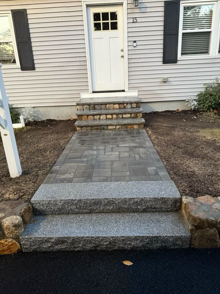 A stone walkway with two granite steps in the foreground leads to the front door stairs on a house with light siding.
